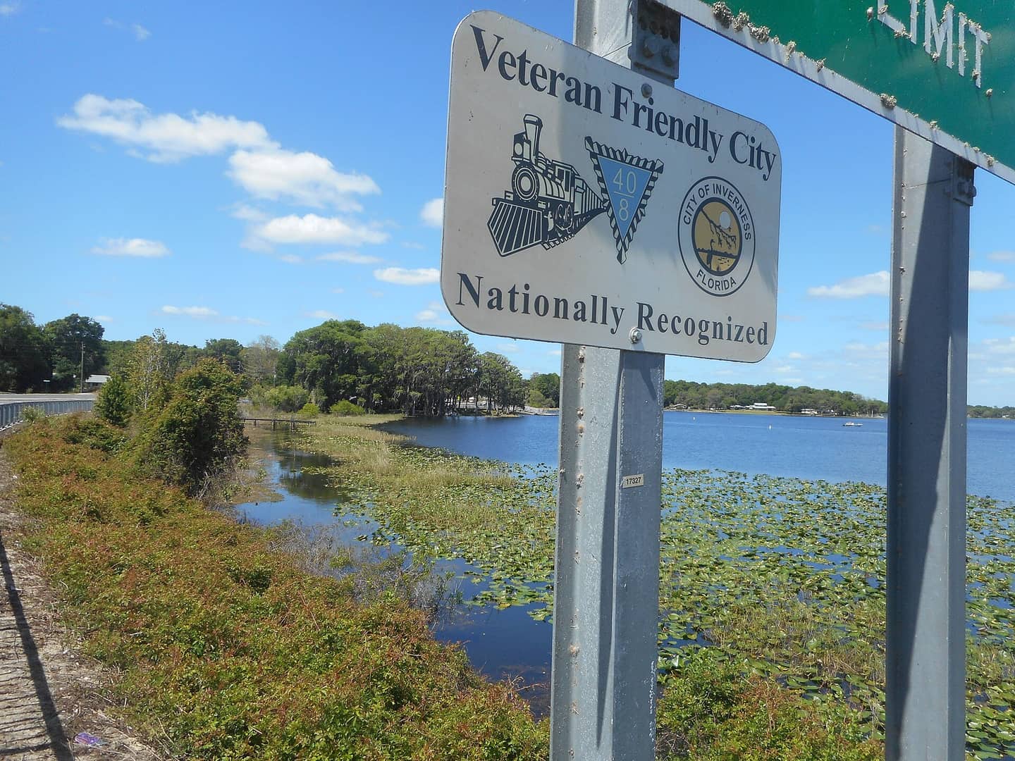 Veteran Friendly City sign beside a calm lake with lily pads and trees under a blue sky.