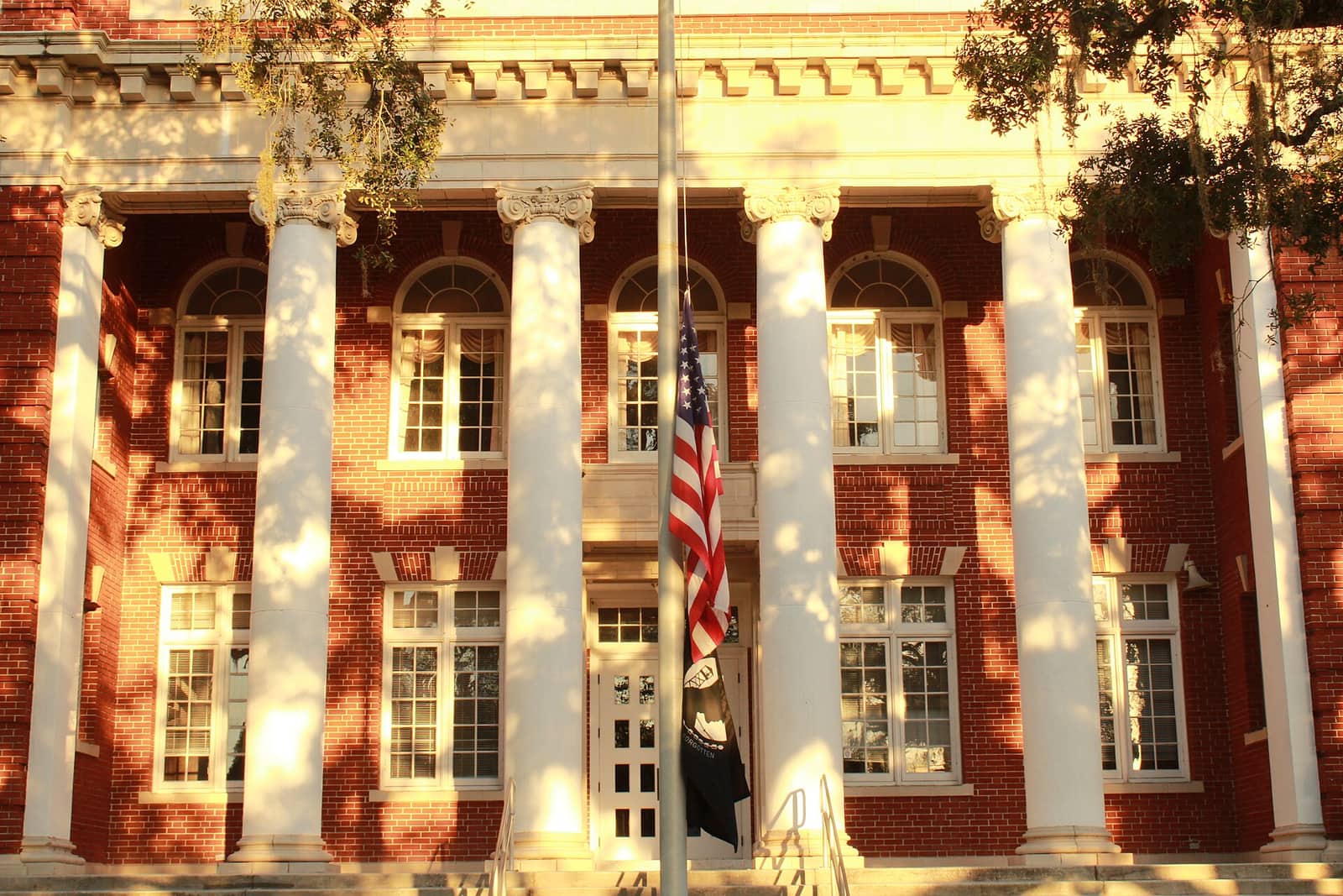 Red brick courthouse facade with white columns, a flag, and dappled sunlight across windows.