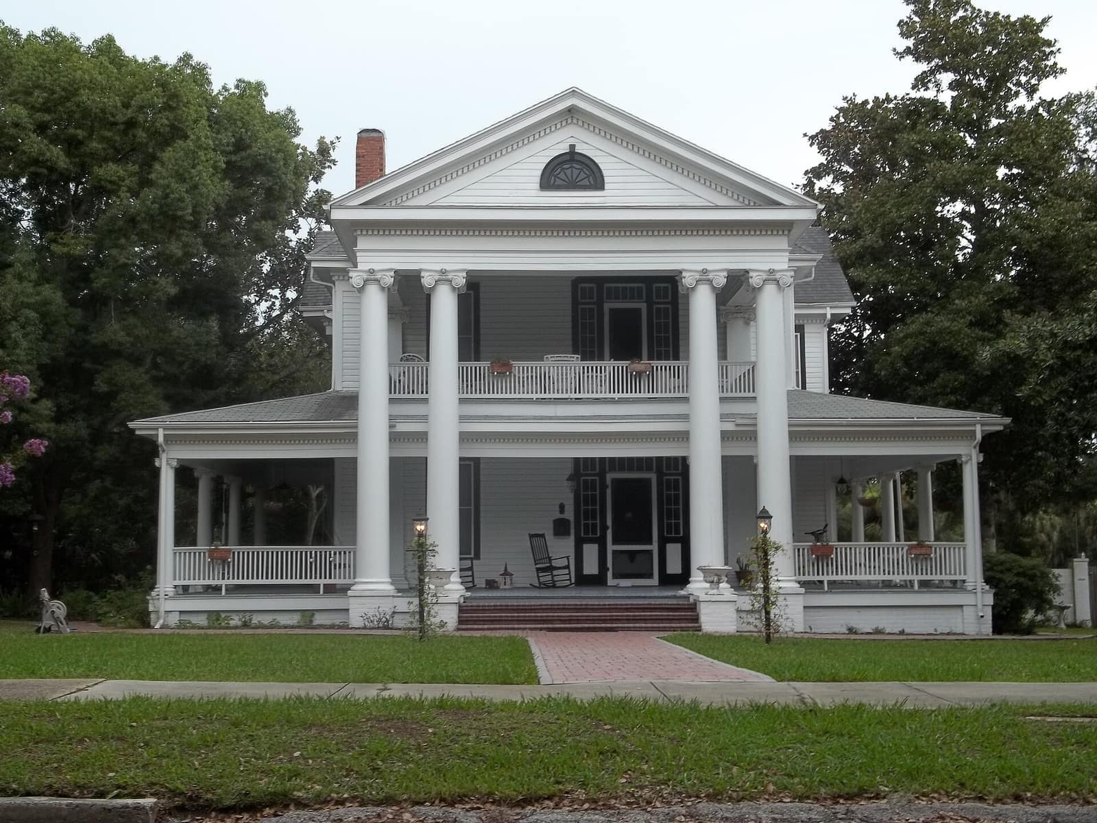 A white two-story columned historic house with a wide front porch and rocking chairs under trees.
