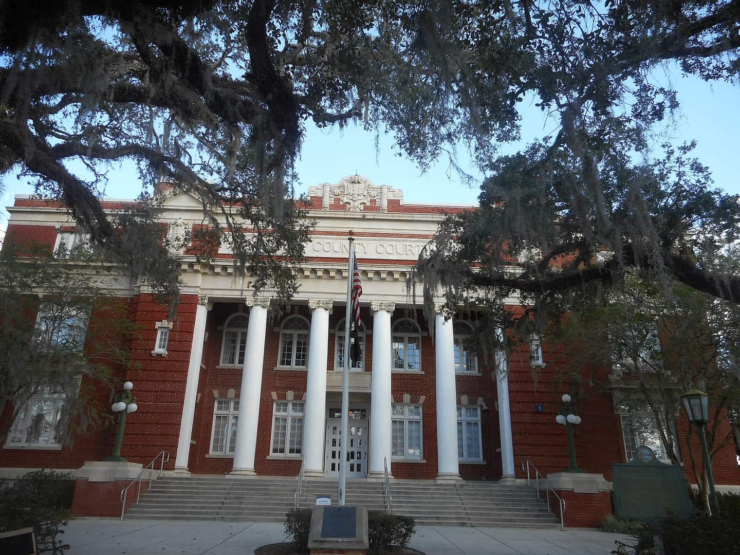 Red brick courthouse with white columns, framed by hanging Spanish moss and oak branches.