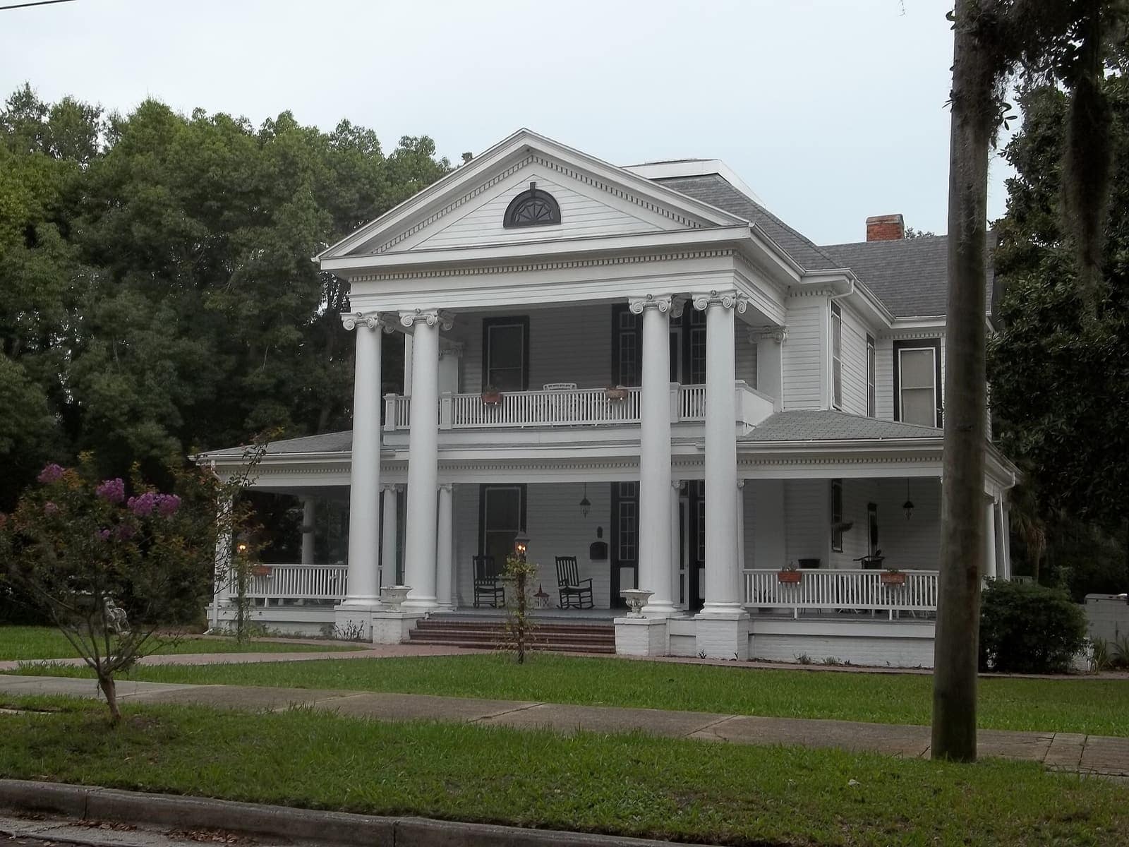 Side-angle view of a white columned historic house with a two-story front porch under tall trees.
