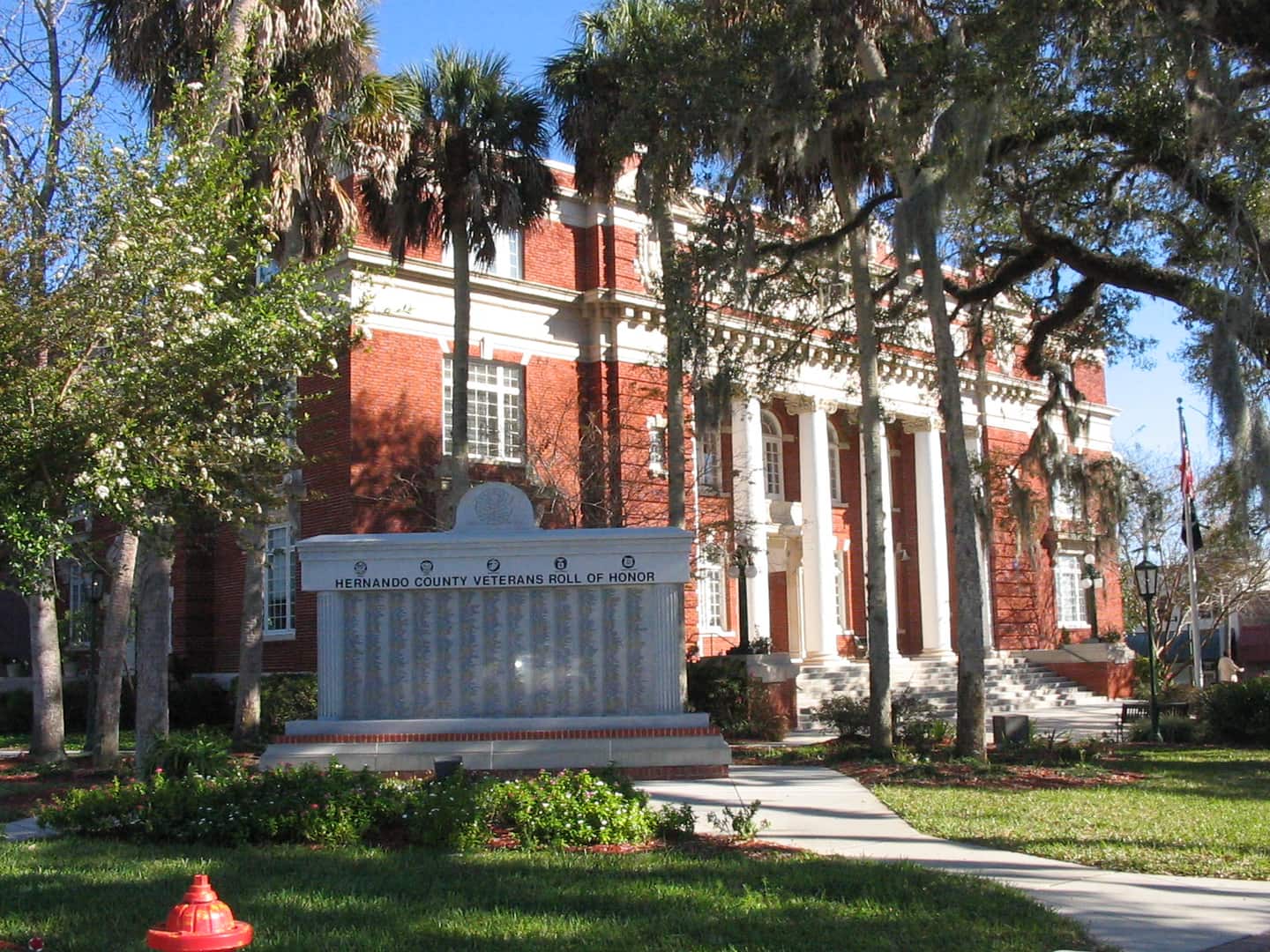 Red brick county courthouse with white columns stands behind a veterans memorial wall and palm trees.