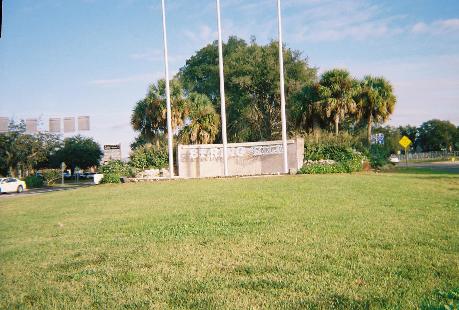 Green lawn in front of a low stone entrance sign with palm trees and flagpoles behind it on a sunny day.