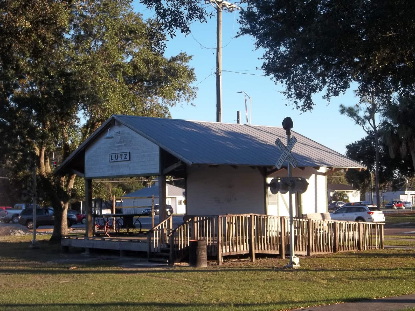 Small open-sided wooden depot shelter with a metal roof, a sign reading Lutz, and a railroad crossbuck.