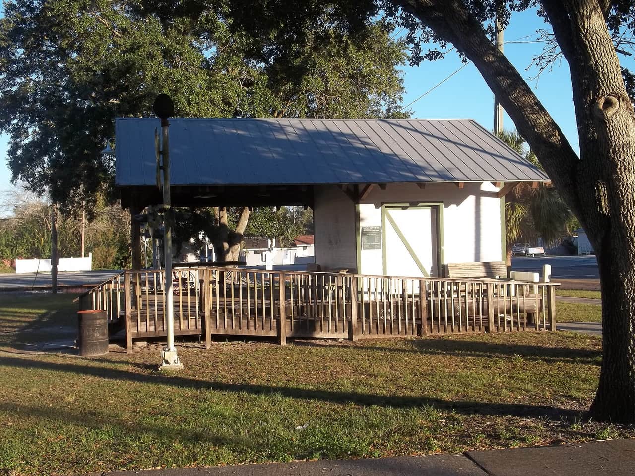 Rebuilt wooden depot shelter with a metal roof and railing, shaded by a large oak tree.