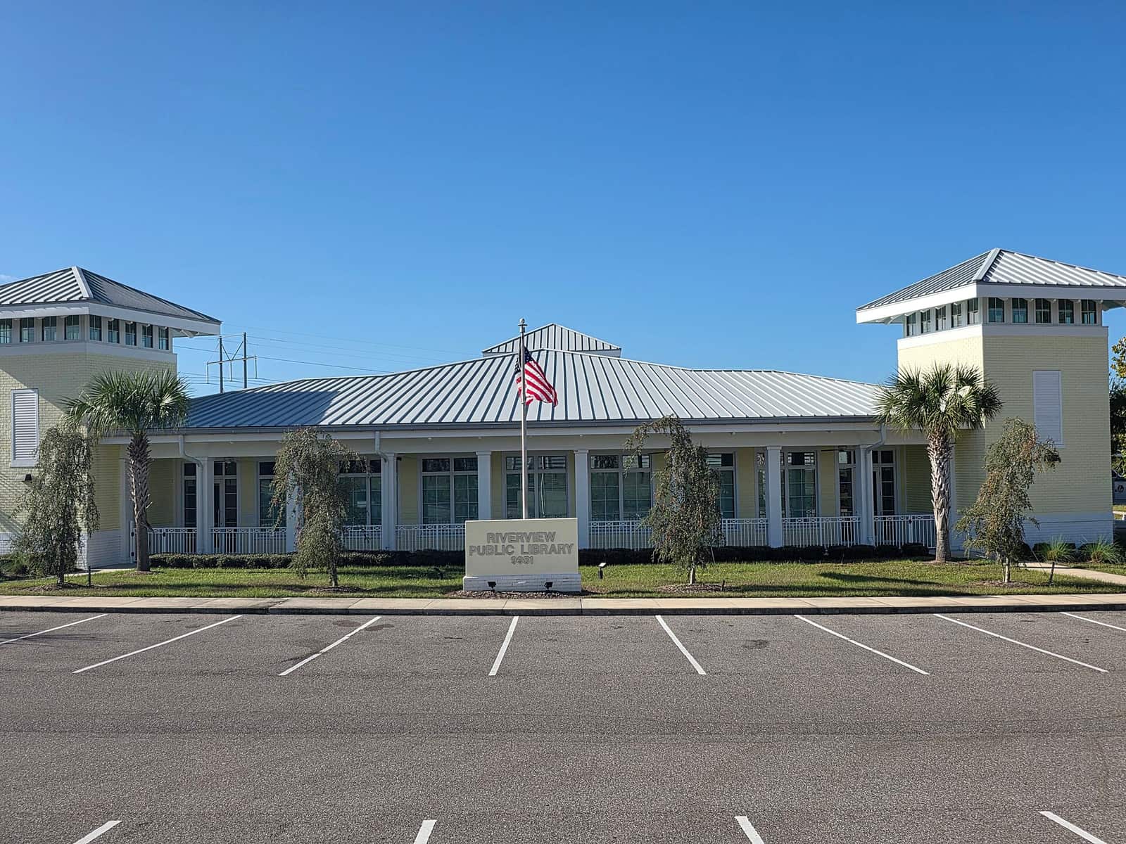 Single-storey yellow public library with metal roof, flagpole, palm trees, and an empty parking lot.