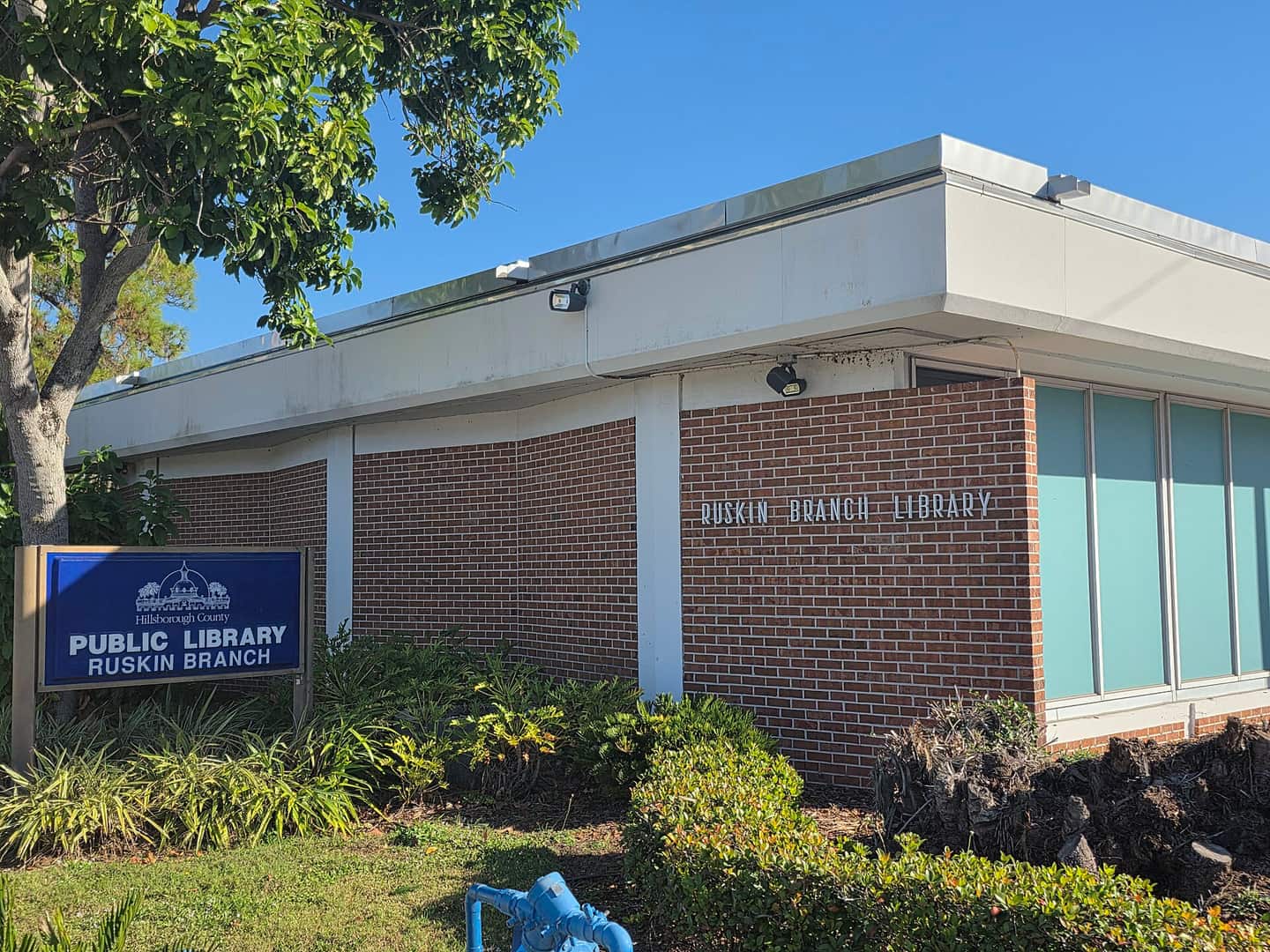 Brick library building with a blue Public Library sign and green shrubs under a clear blue sky.