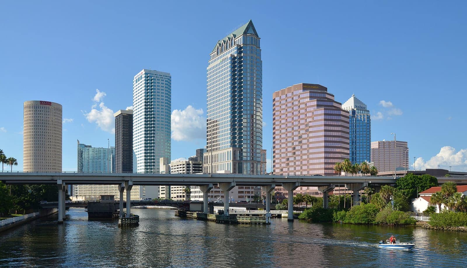 Downtown high-rise towers rise above a river with a elevated bridge and a small boat on the water.