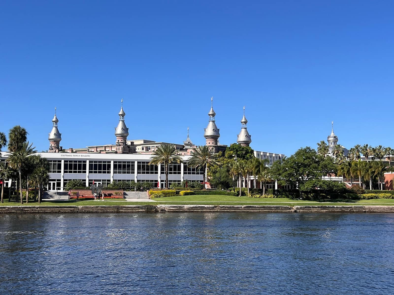 A large ornate building with several domed turrets stands behind a riverside lawn and palm trees under a clear blue sky.