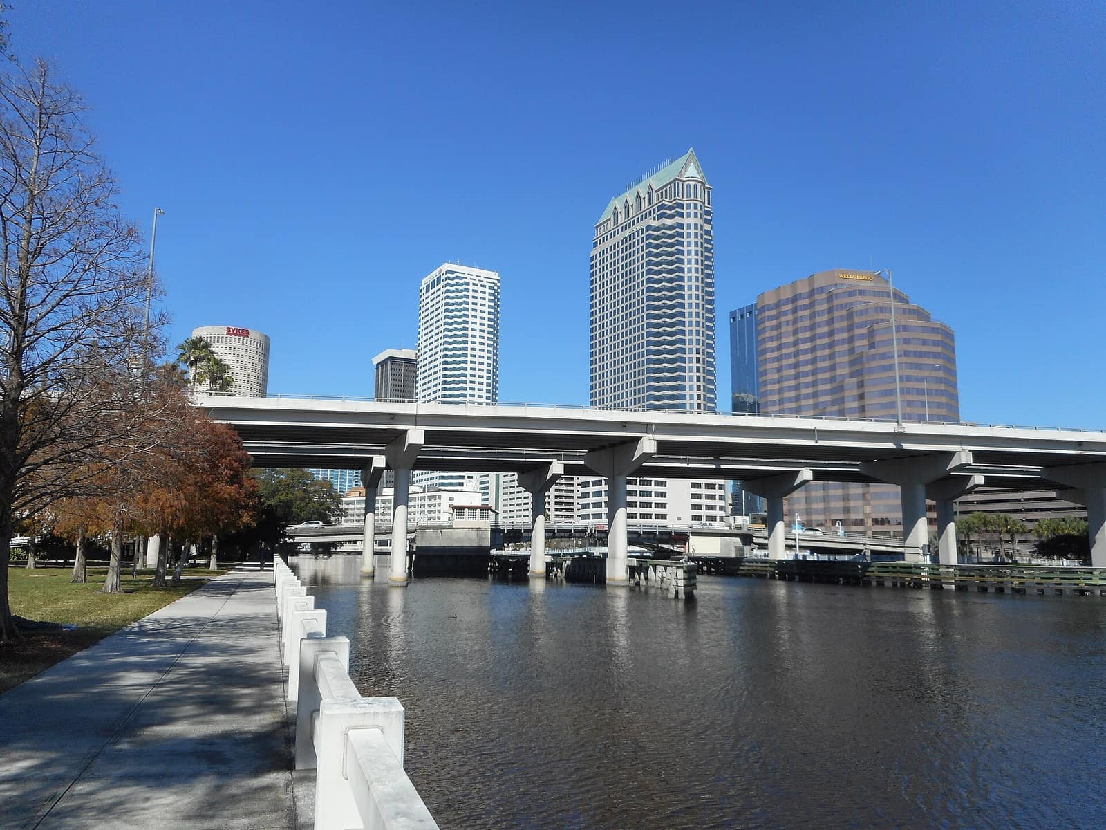 A wide concrete elevated highway crosses a river with downtown office towers behind it under a clear blue sky.