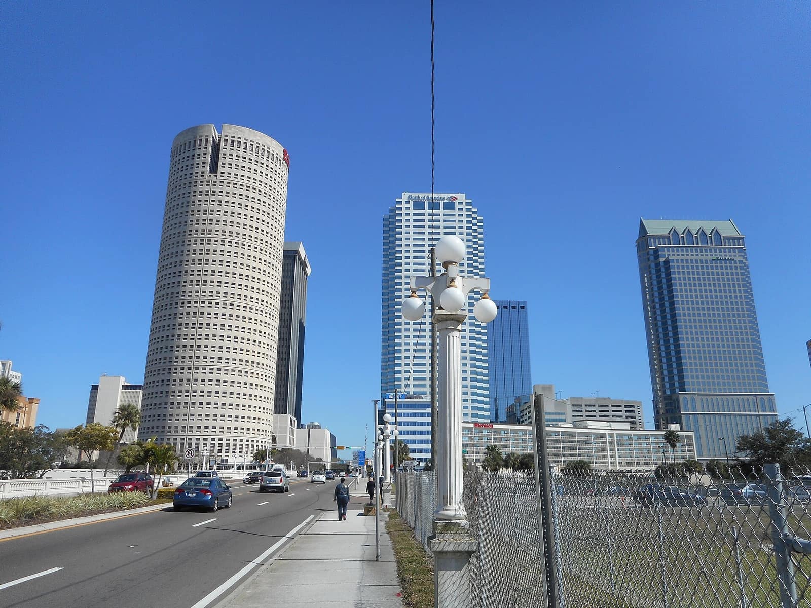 Downtown office towers beyond a decorative lamp post, sidewalk, fence and a road under blue sky.