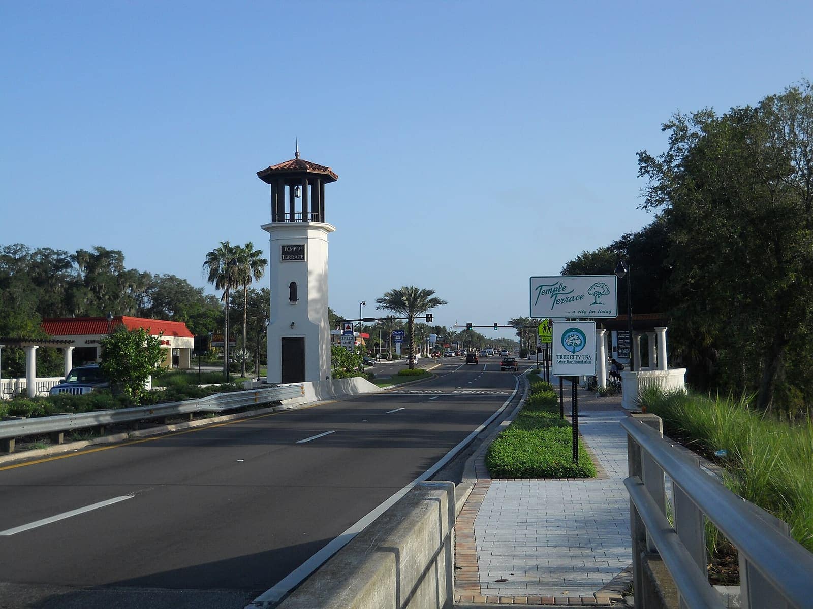 A tall white tower with a red-roofed cupola stands beside a divided road lined with palm trees.