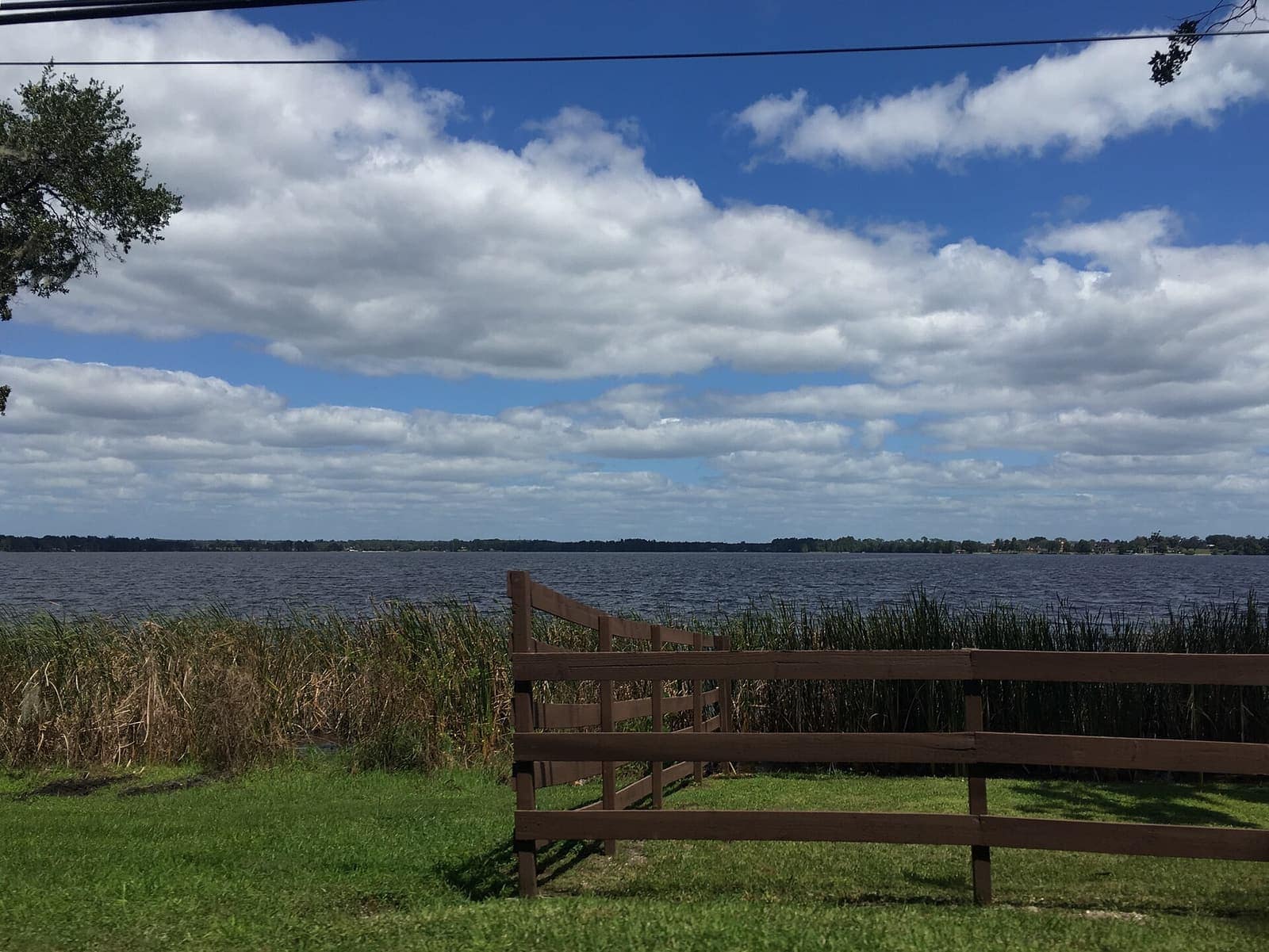 Wooden fence in grass overlooks a wide lake under large white clouds and a power line.