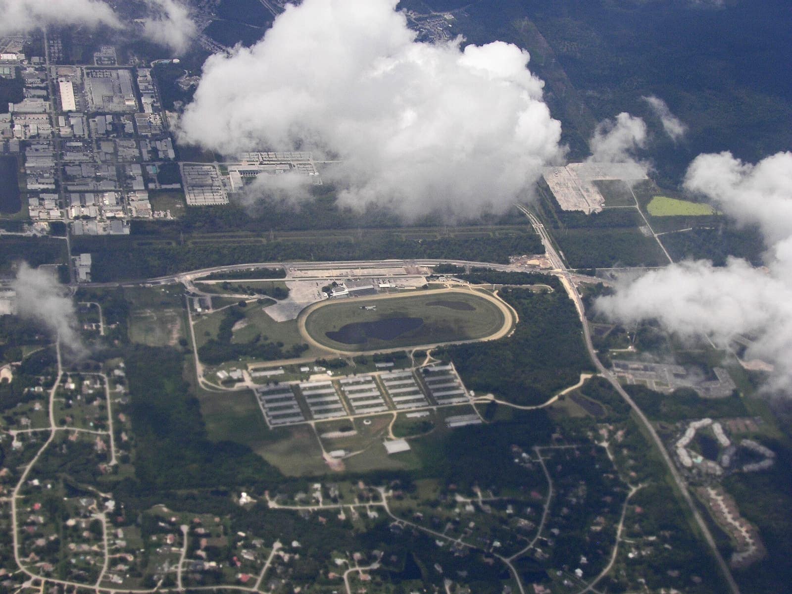 Aerial view of an oval racetrack with a grandstand, low rows of buildings and surrounding wooded land.