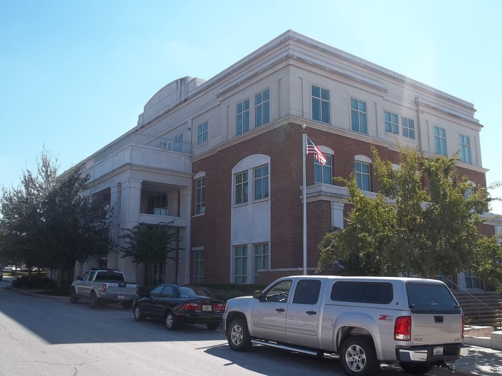 Three-story brick and cream civic building with an American flag beside parked pickup trucks and cars.