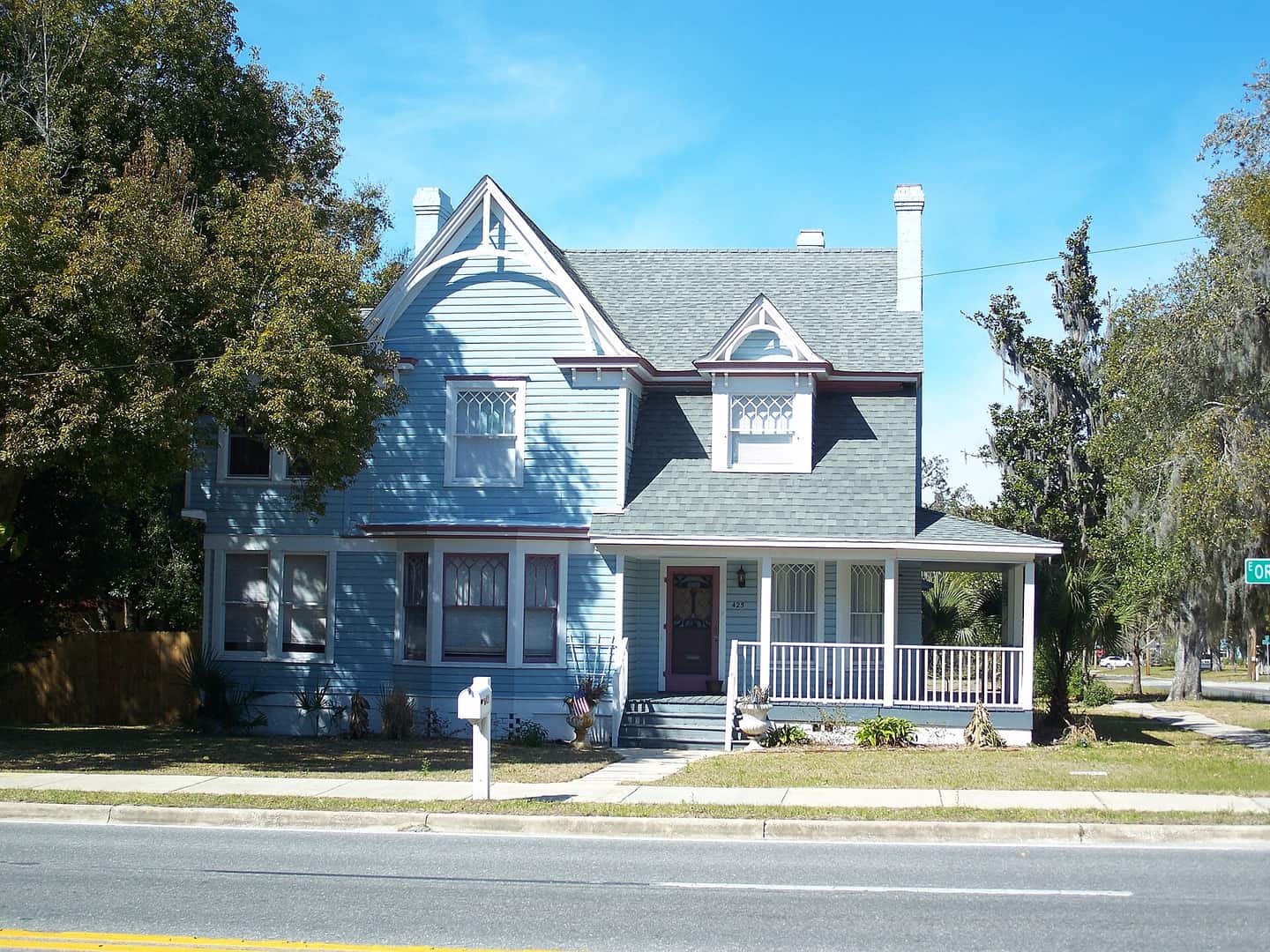 Blue Victorian two-story house with a front porch, gabled roof, and tall oak trees under blue sky.