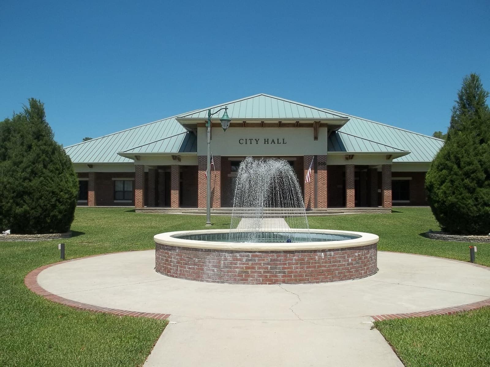 Single-storey brick city hall with teal metal roof, fountain, and lawn under clear blue sky.