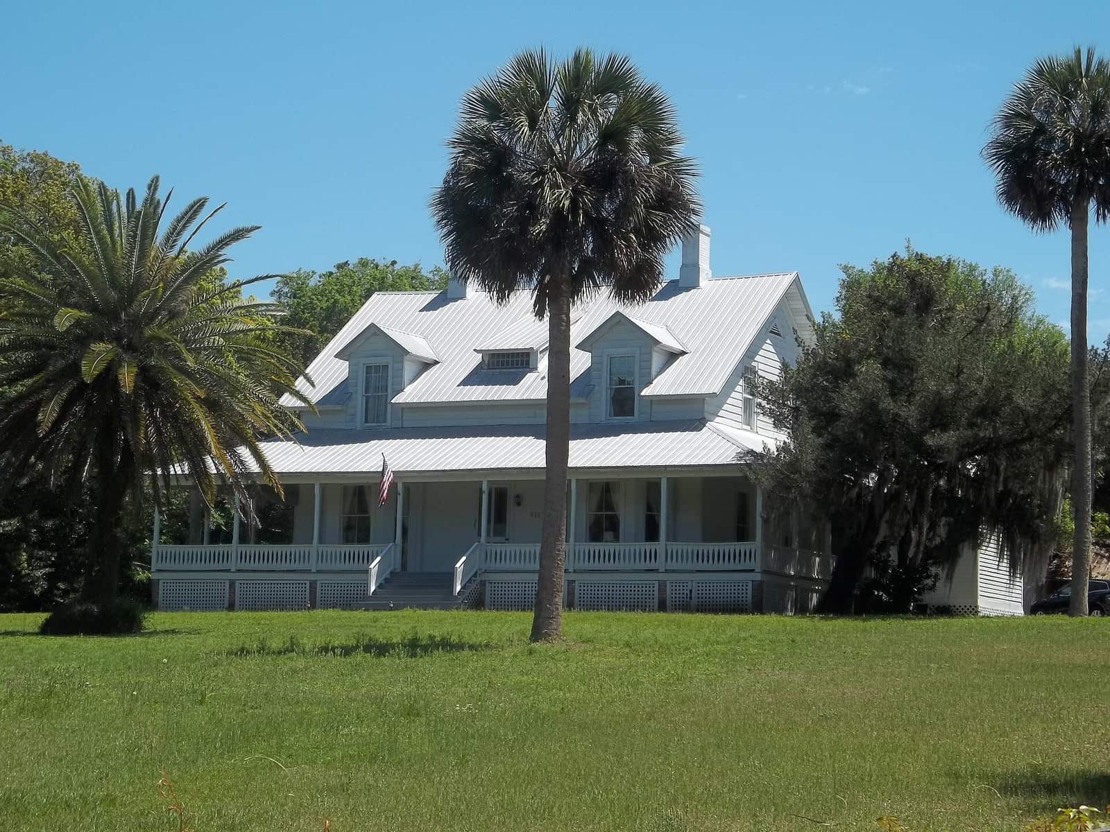 White two-storey wooden house with wraparound porch and palm trees on green lawn.