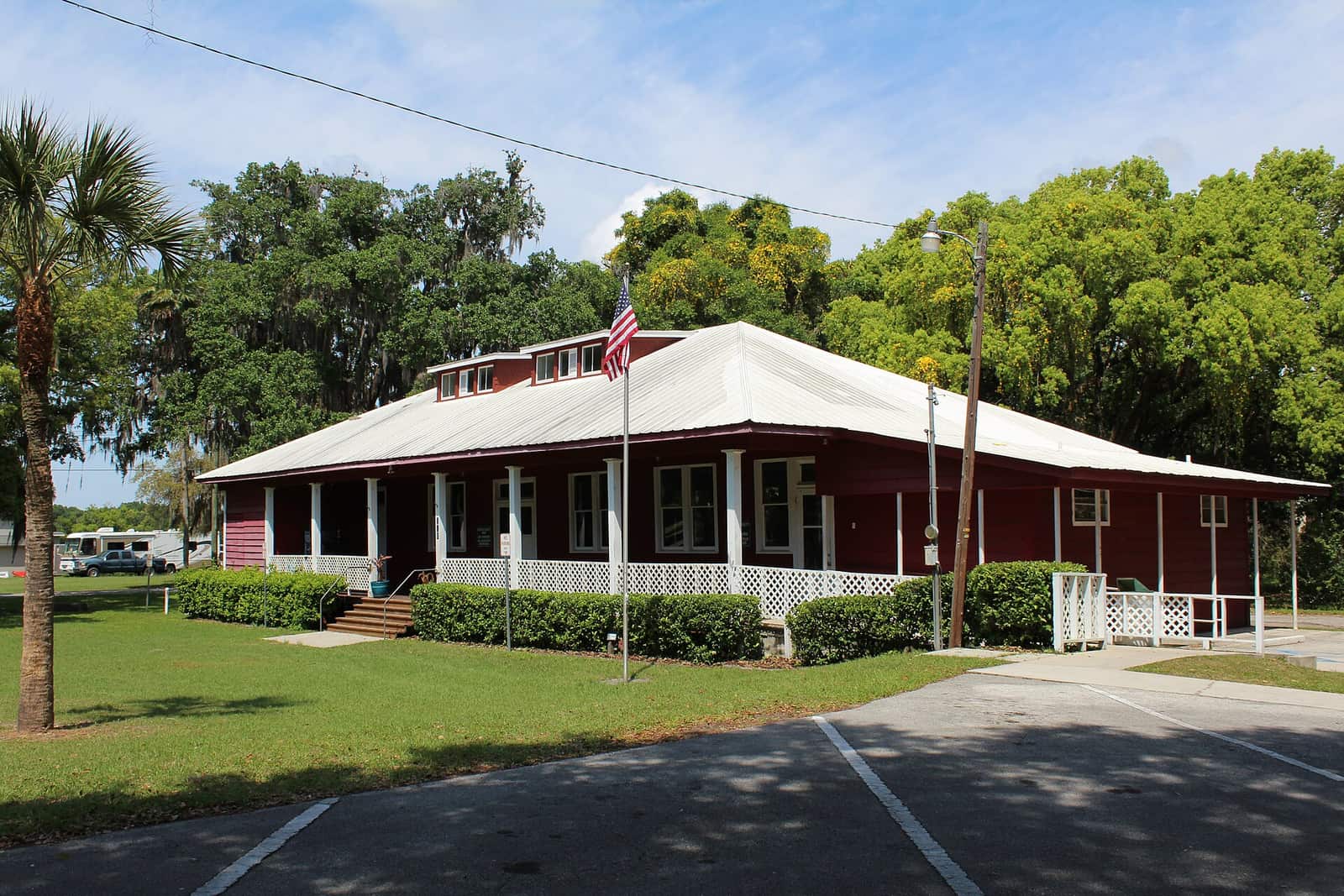 A red wooden community building with white porch railings, a metal roof, and an American flag, shaded by trees.