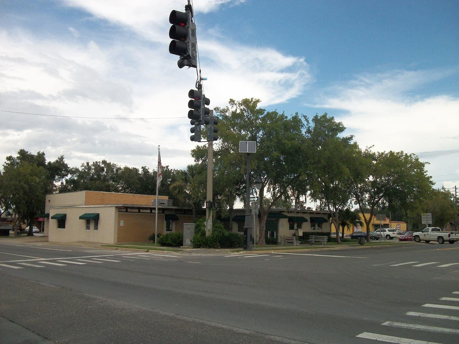 A small beige city hall building with green awnings beside traffic signals and parked cars under a cloudy sky.