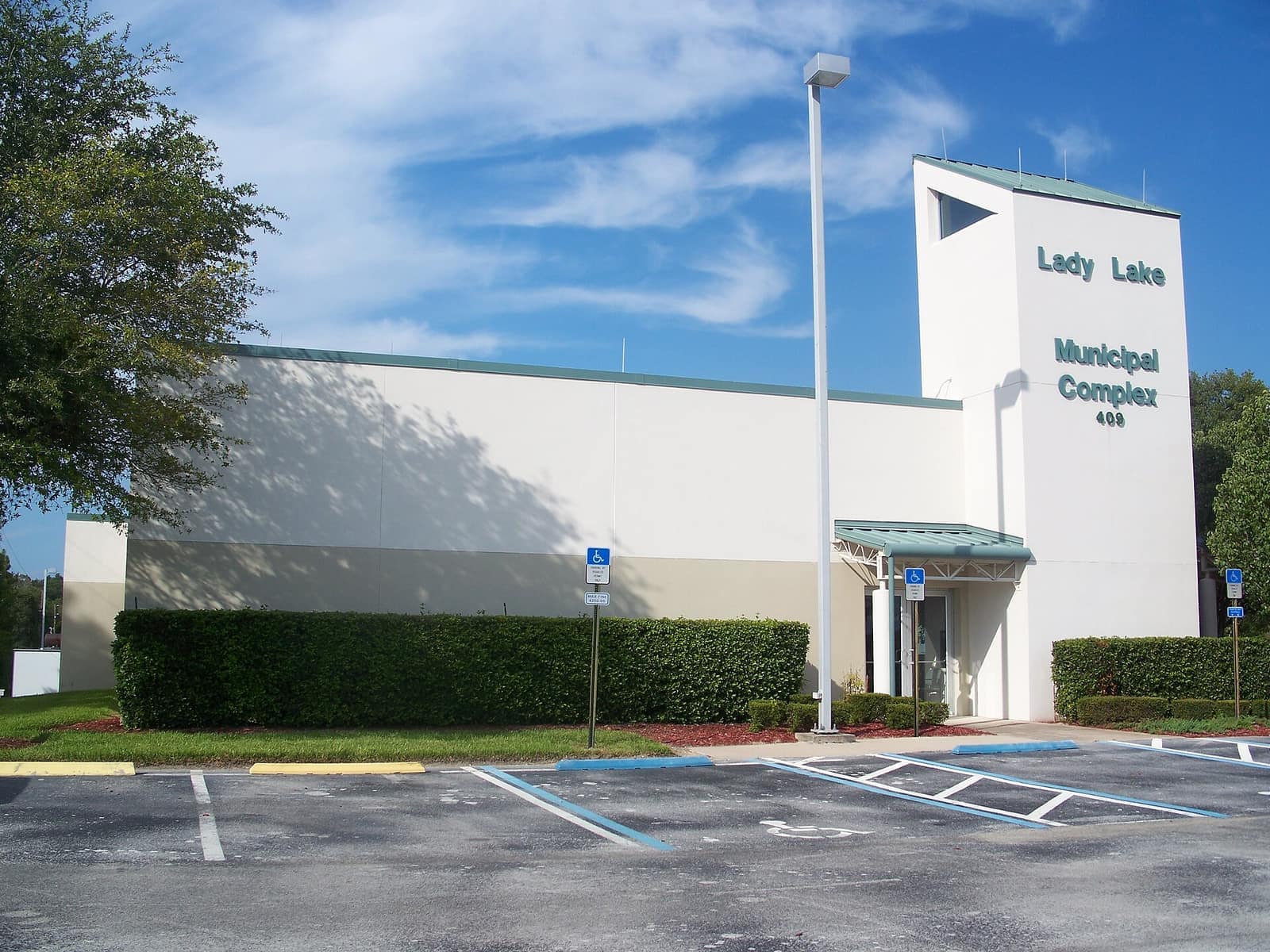 Modern white municipal complex building with a tower and entrance canopy, shrubs and parking spaces in front.