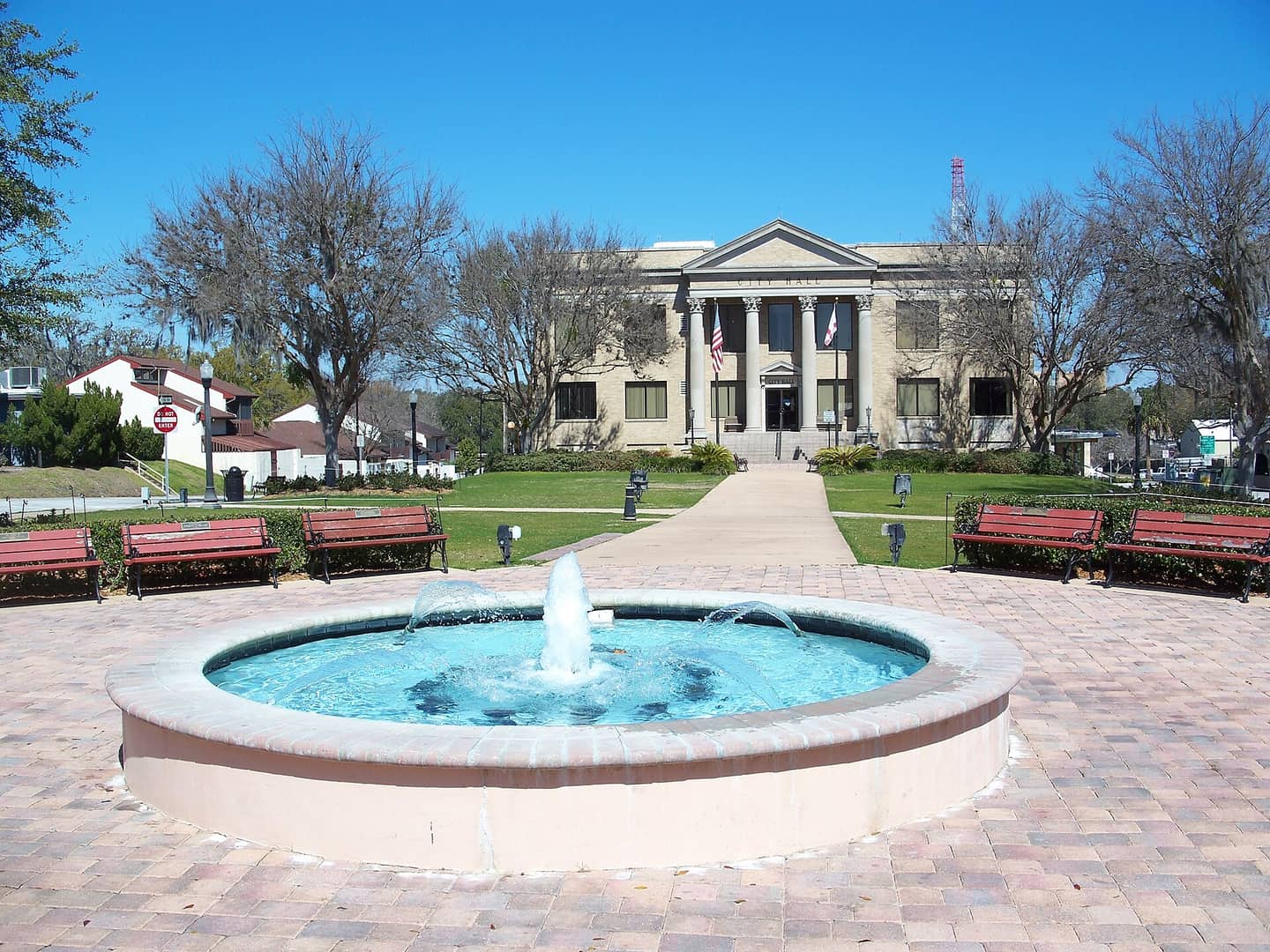Neoclassical city hall behind a round fountain and red benches under a clear blue sky.