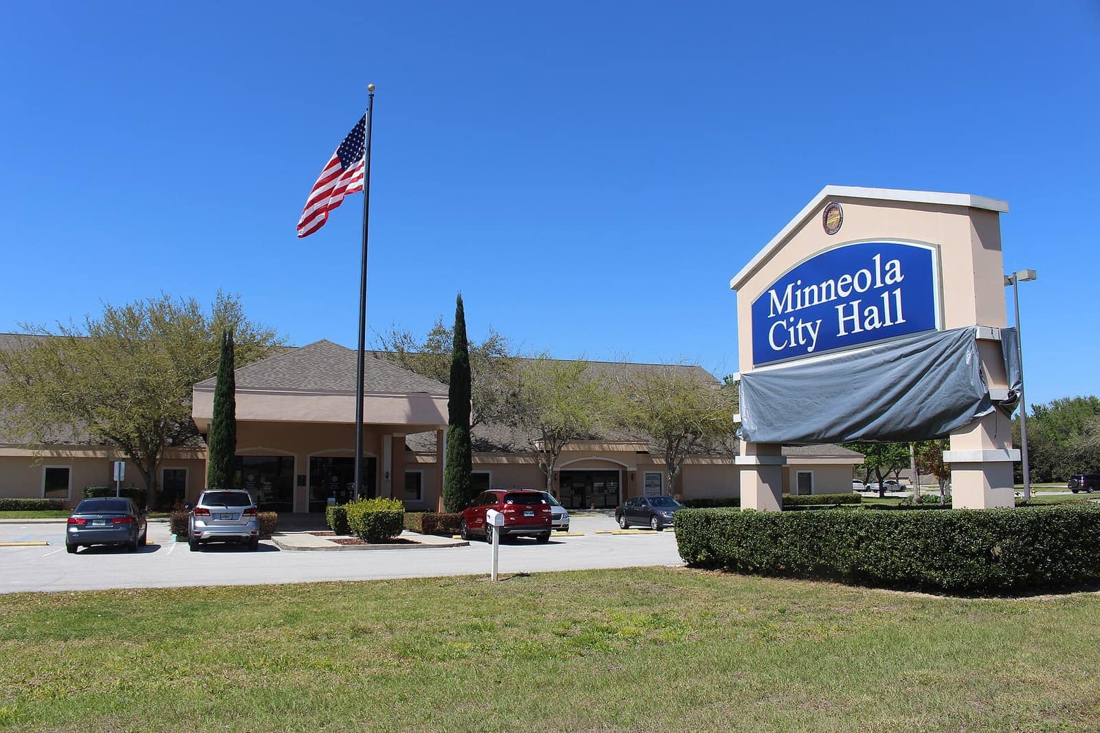 Minneola City Hall sign and low tan building beneath a blue sky, with an American flag on a pole.