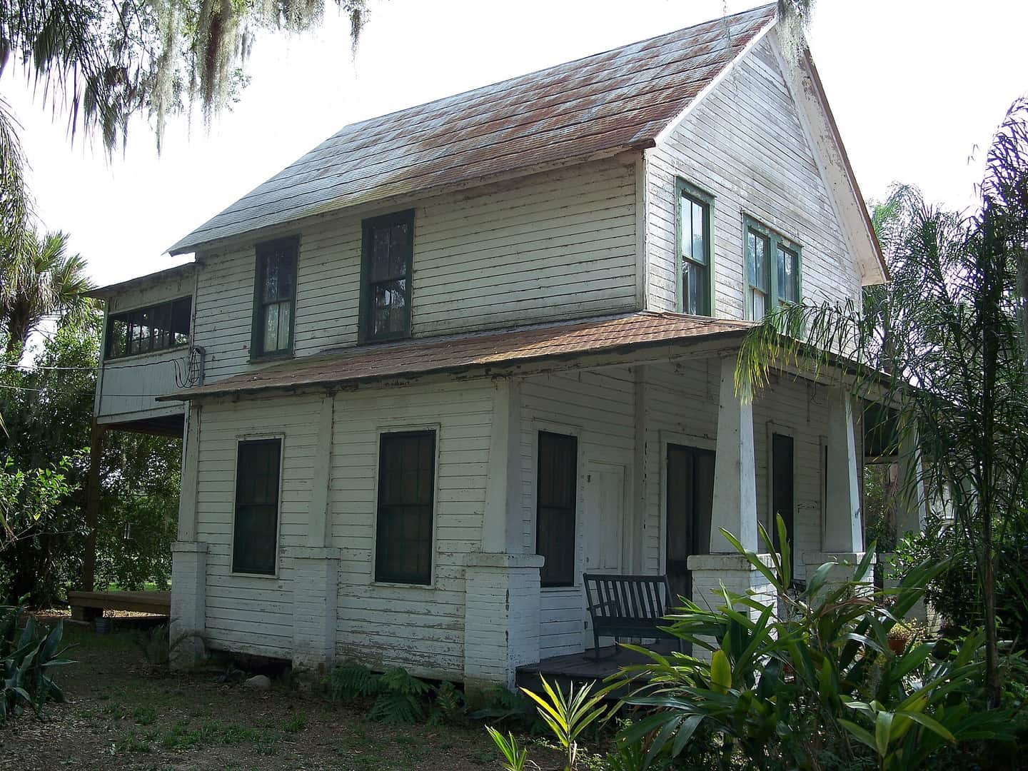 Two-story white wooden house with a front porch, a bench, and tropical plants in the yard.