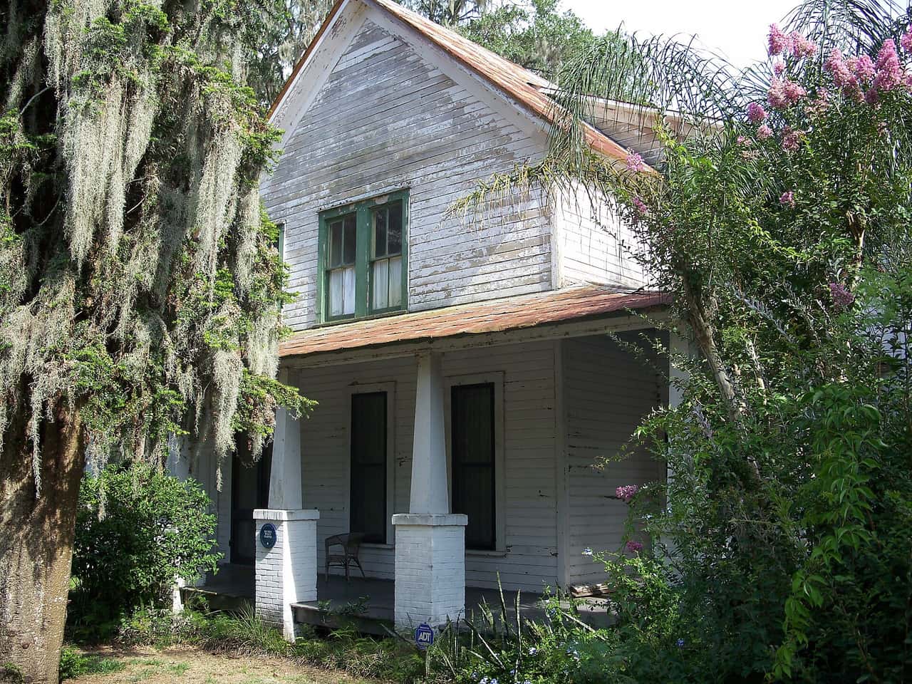 Small white wooden cottage with a front porch, green shutters, and pink flowering shrubs.