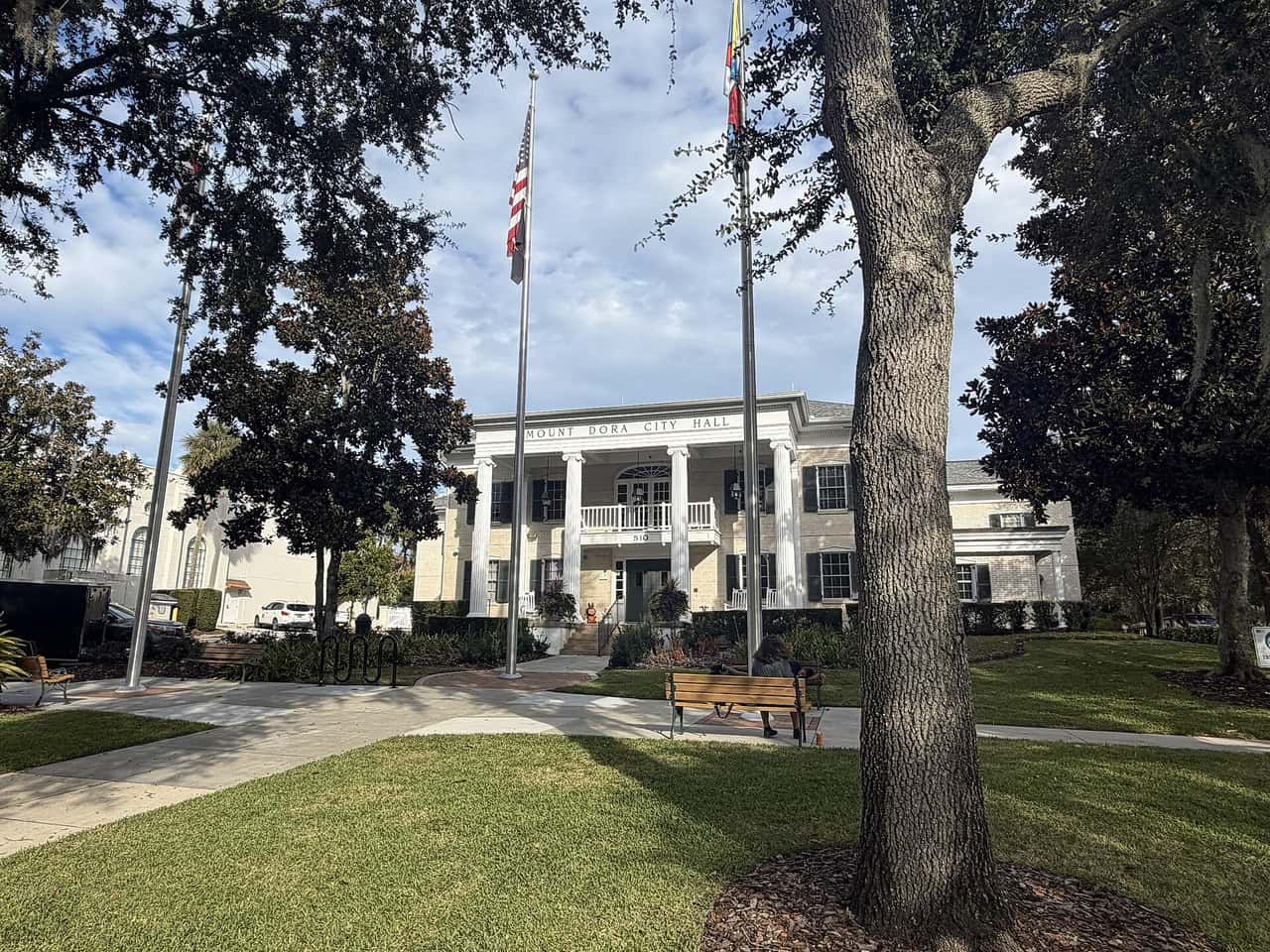 White columned city hall behind a lawn, with two flagpoles, a park bench, and large oak trees.