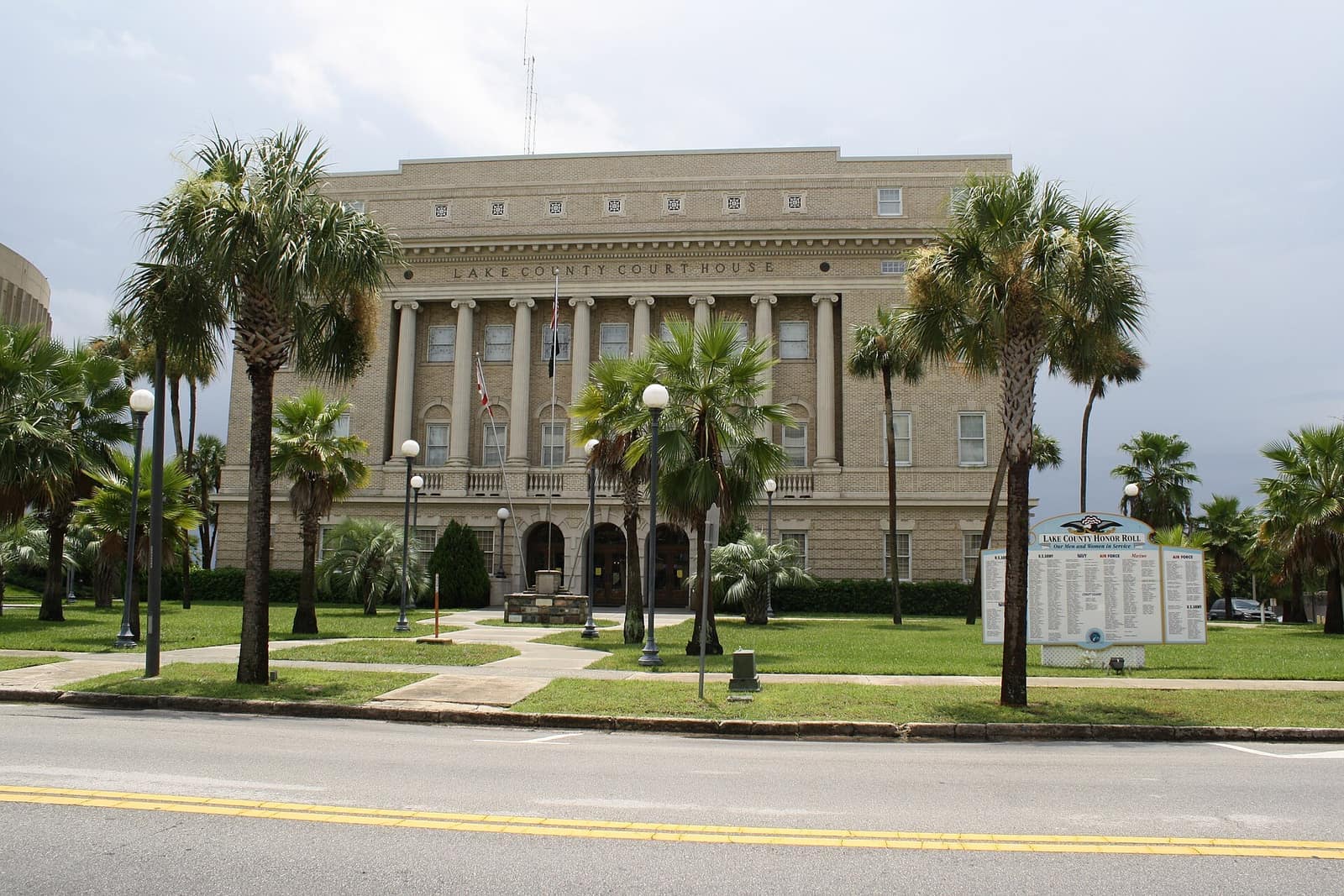Stone courthouse facade framed by palm trees and lawns, with a sign and parked road in front.