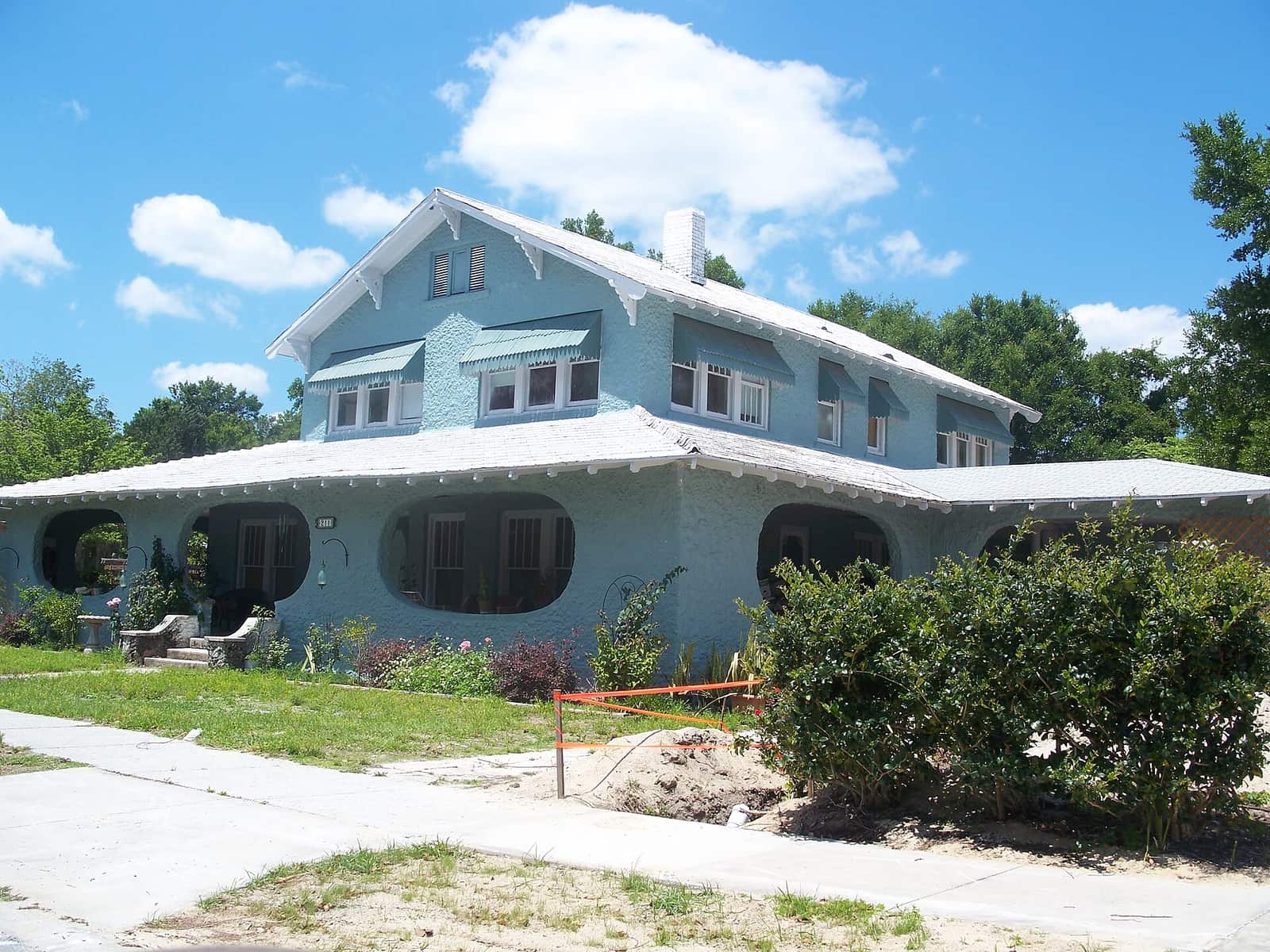 Large light blue two-story house with wraparound porch and bushes under a blue sky.