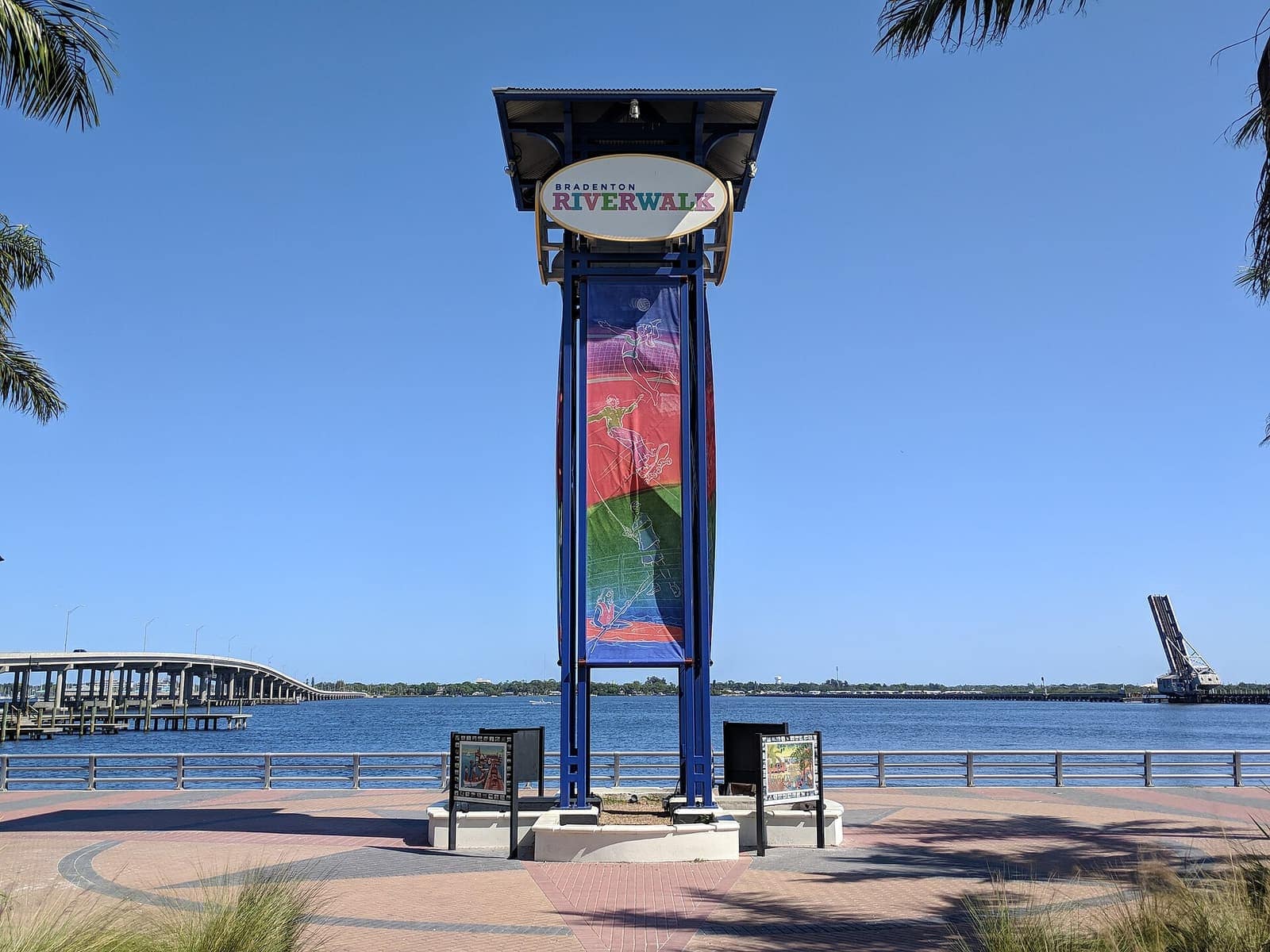 Blue Bradenton Riverwalk tower sign beside a river with a bridge and palm trees under clear blue sky.