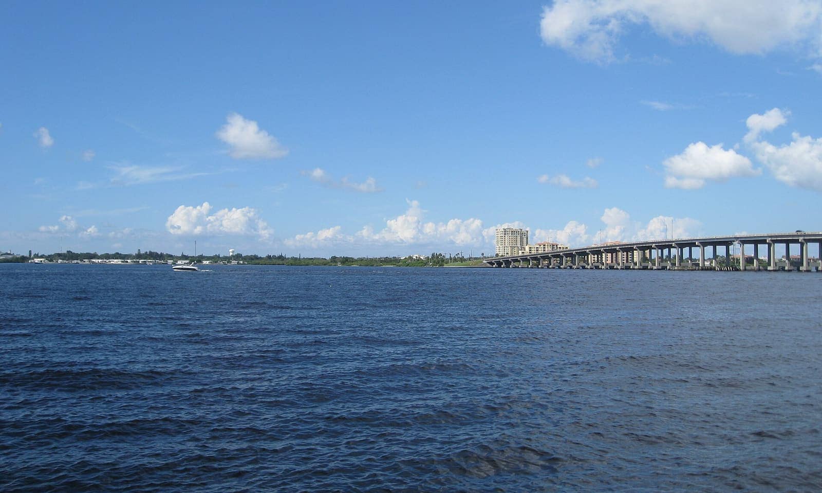 Wide river view with a long concrete bridge on the right, a small boat, and scattered clouds overhead.