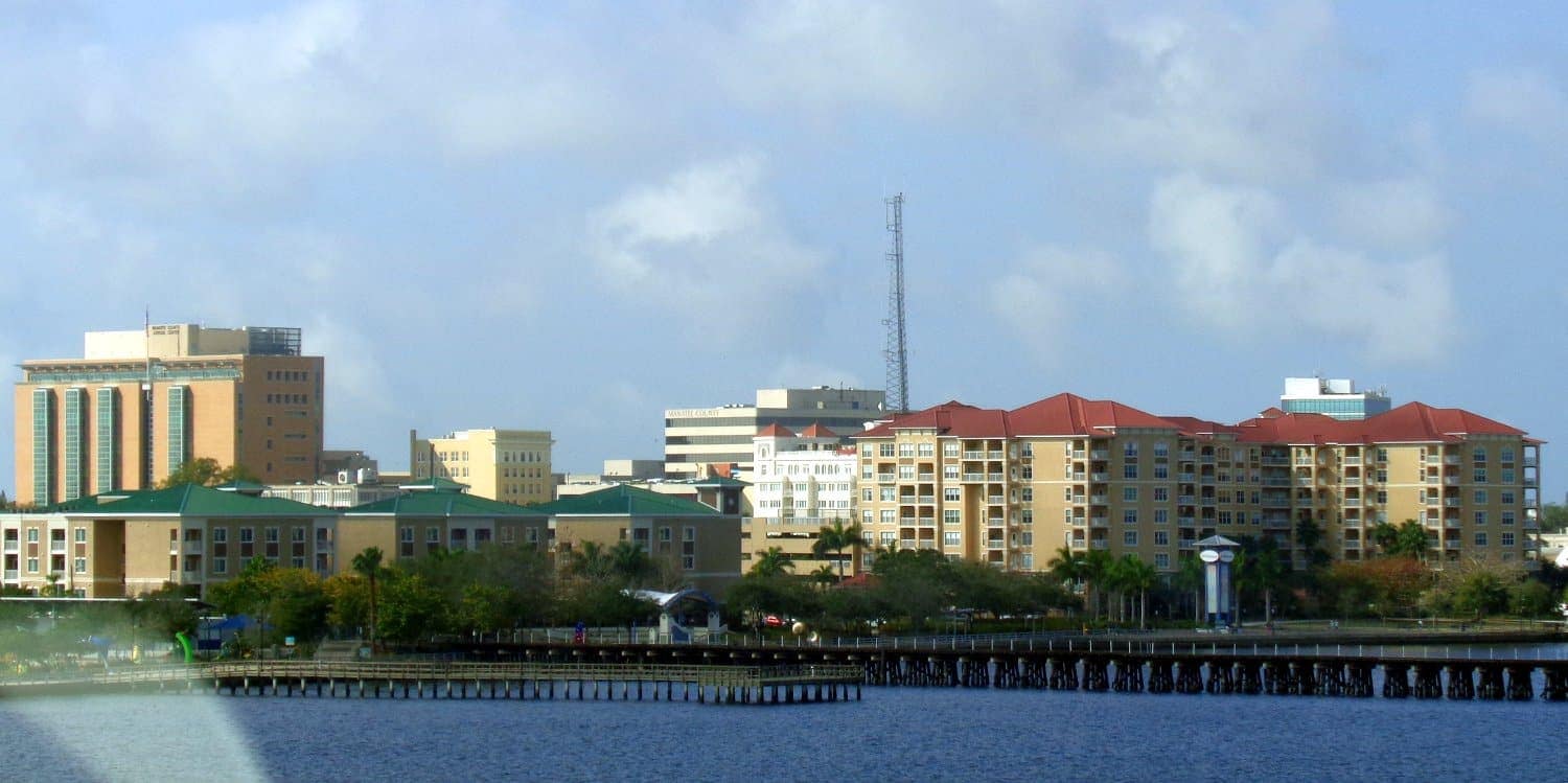 Low-rise and mid-rise buildings rise above a waterfront with a pier and a tall antenna under a cloudy sky.