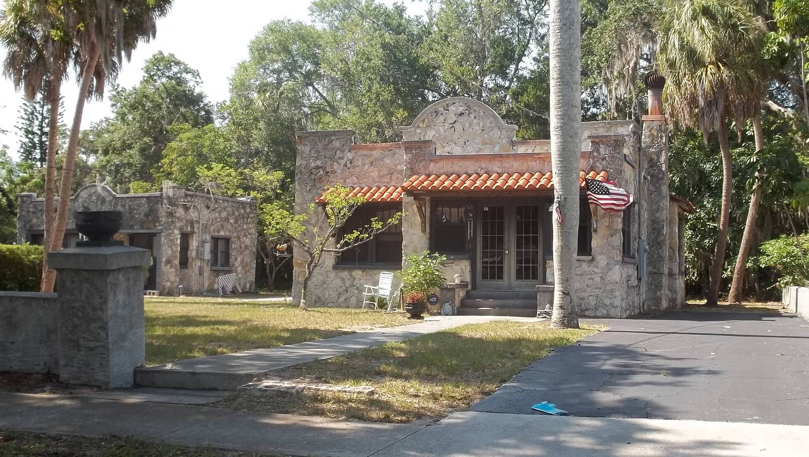 Small stone cottage with a red tile roof, American flag, palm trees, and a stone gateway in the foreground.
