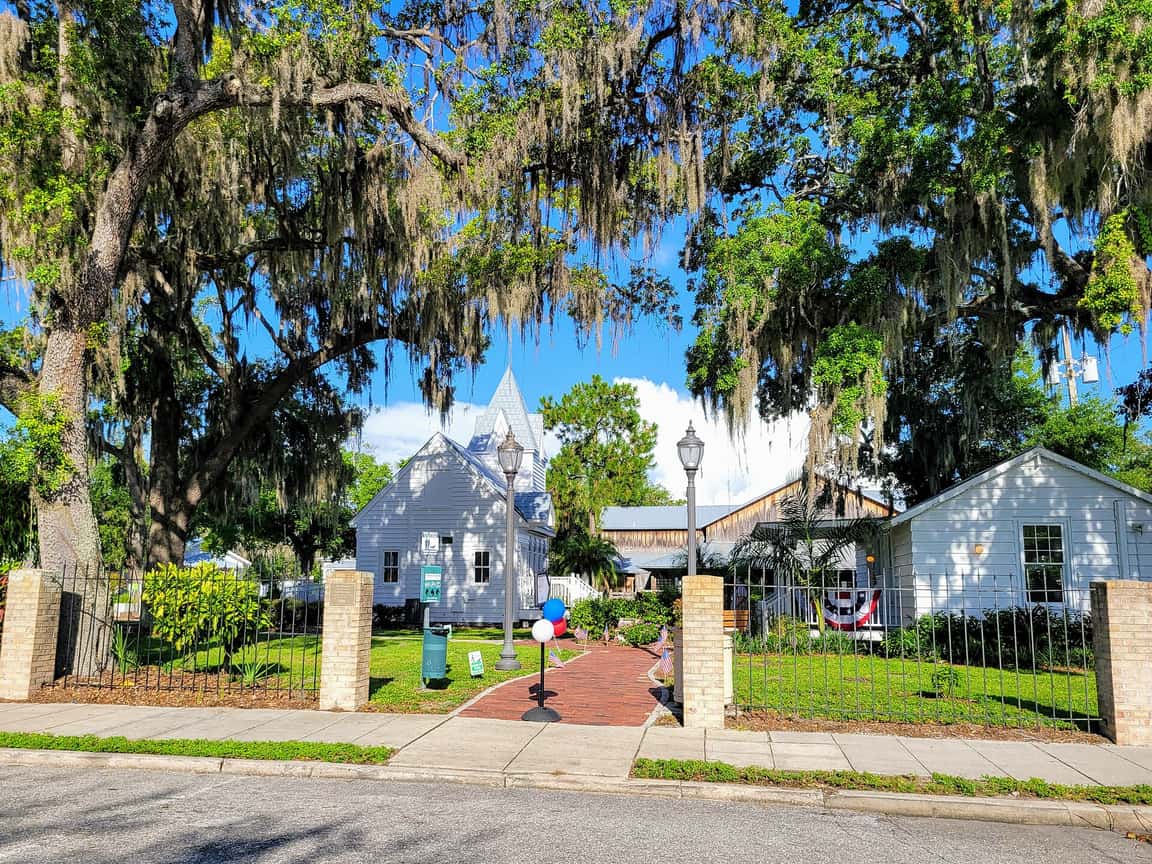 White historic buildings and a brick path under large oaks draped with Spanish moss, seen through a wrought-iron fence.