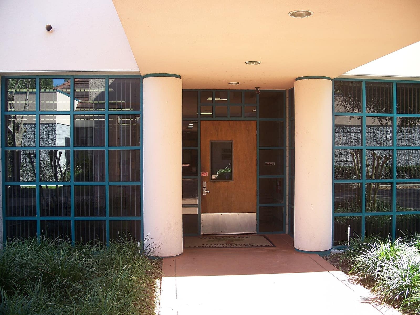 Glass-walled entrance of a modern building with cylindrical columns and a wooden door in daylight.