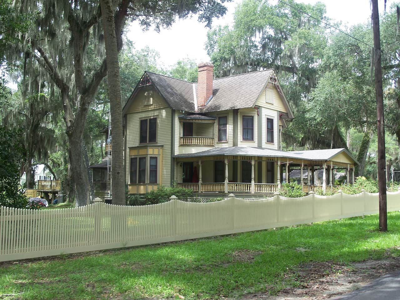 Two-story yellow Victorian house with a wraparound porch behind a pale picket fence.