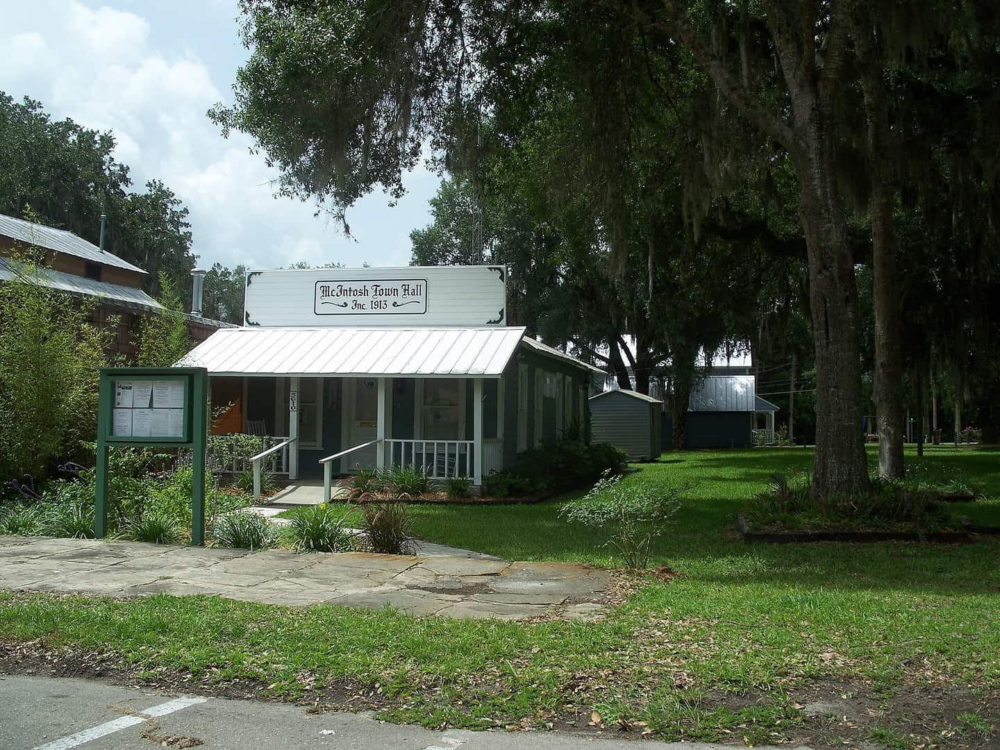Small white town hall with a metal roof and front porch, shaded by large oak trees.