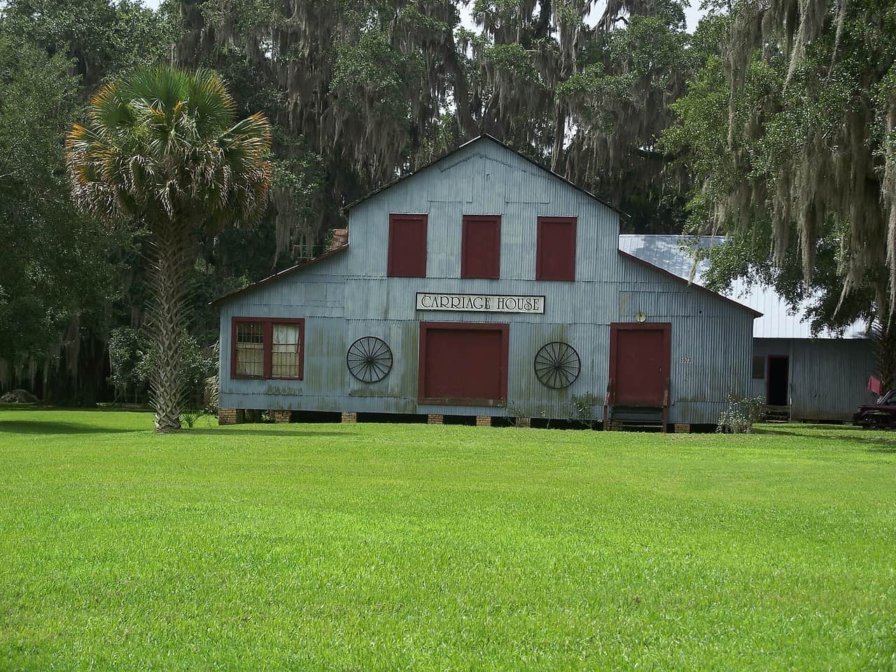 Weathered gray wooden building with red doors and wagon wheels on its front, beside a palm tree.