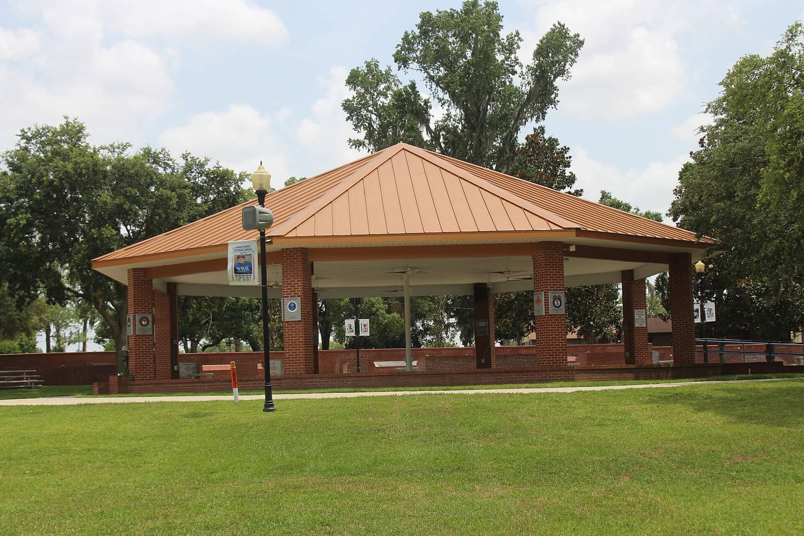 Brick pavilion with copper metal roof stands on green lawn beside tall shade trees in daylight.