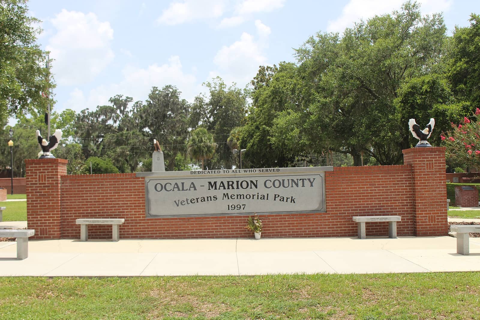 Brick entrance wall with engraved park name plaque, stone benches and eagle sculptures under a blue sky.