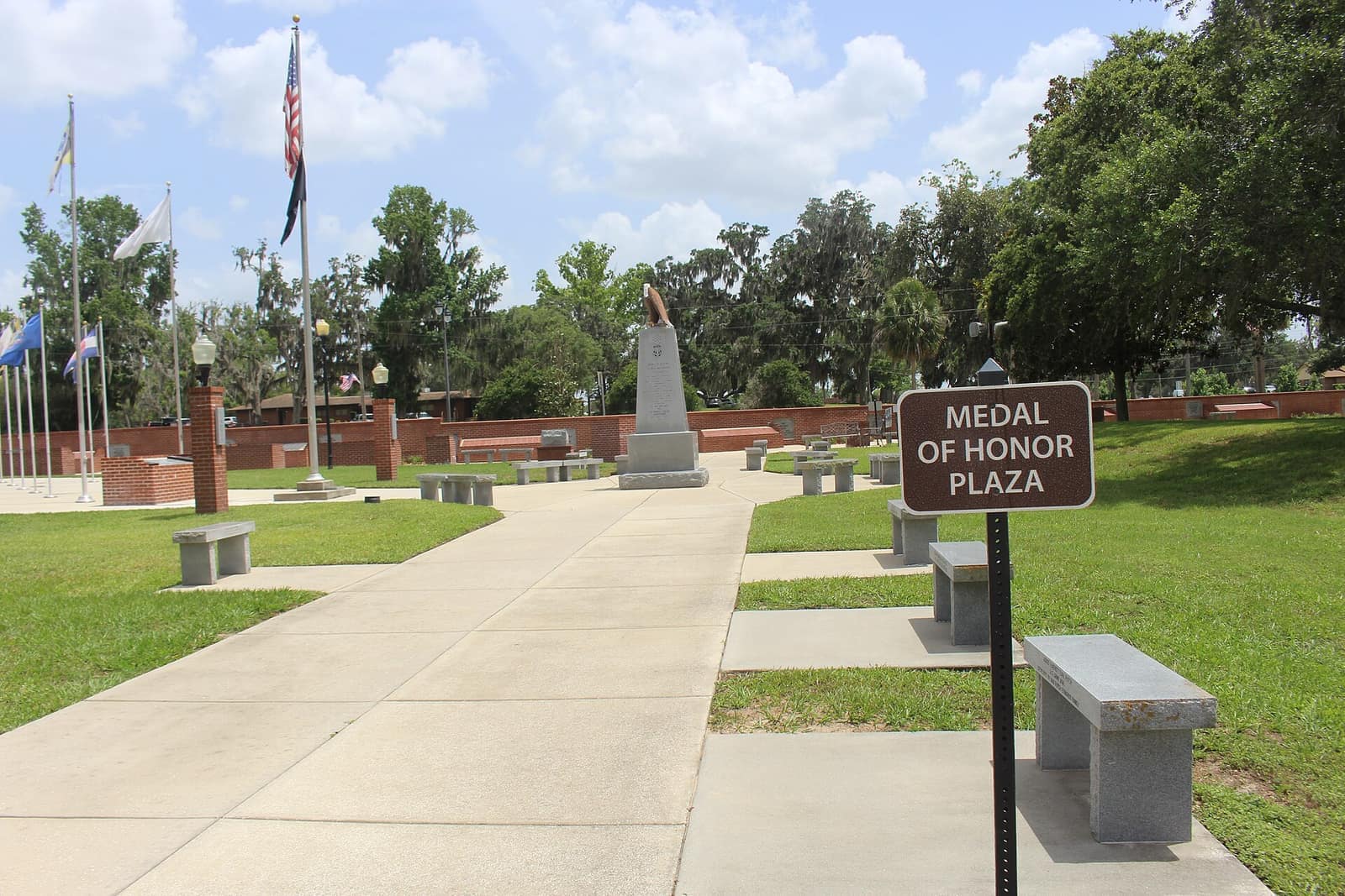 Concrete walkway leads past a memorial monument, flagpoles and benches toward a brick wall under blue sky.