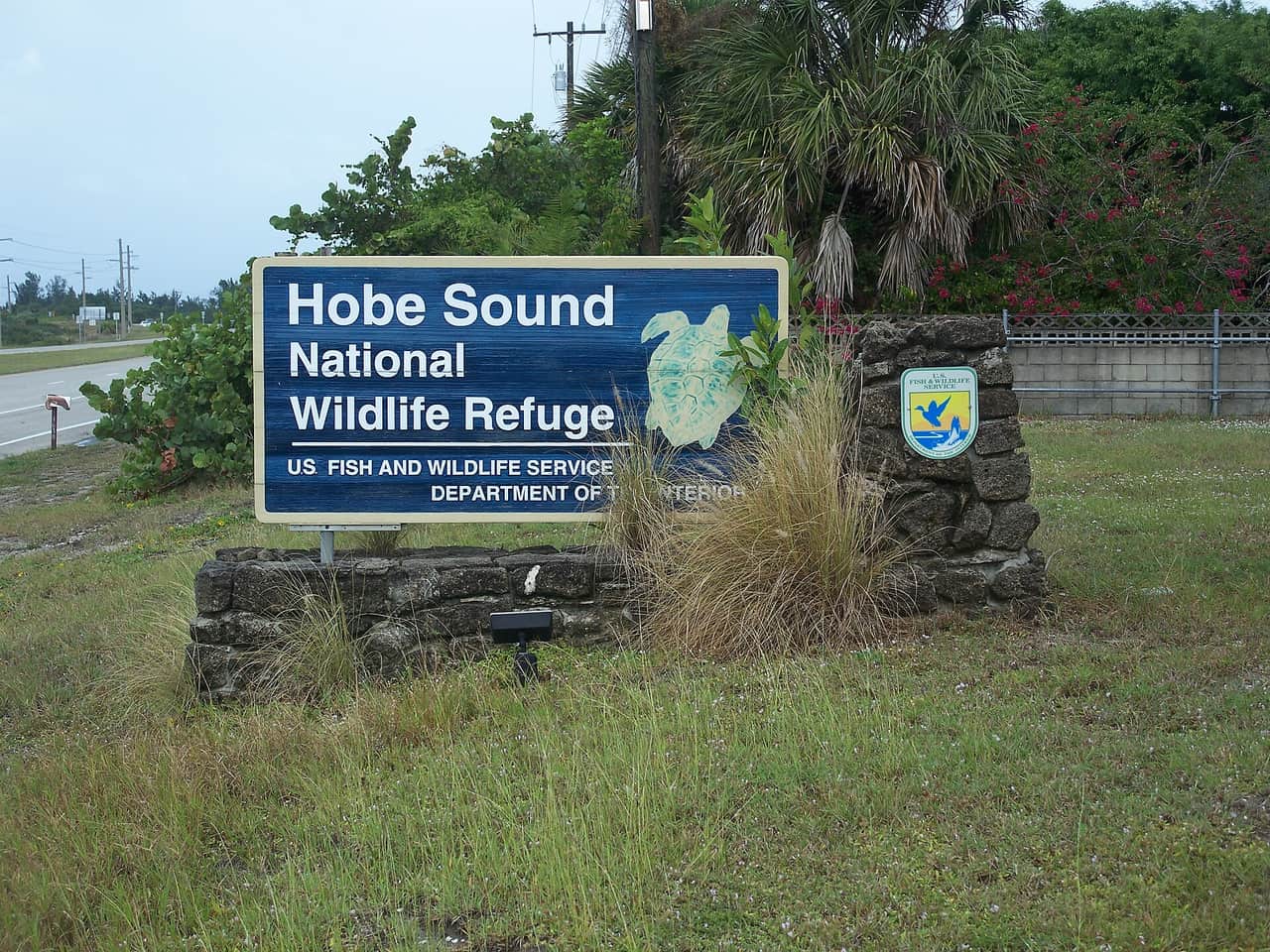 A blue Hobe Sound National Wildlife Refuge sign with a sea turtle graphic on a stone base in grass.