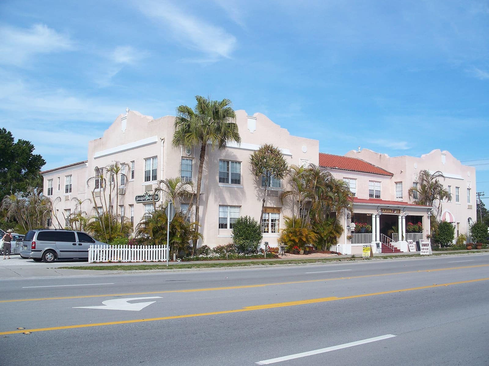 Pink stucco multi-story historic inn with arched parapets and palm trees beside a wide road.