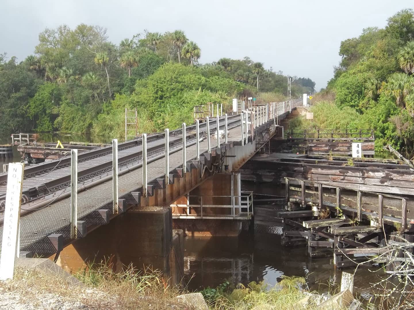 Old single-track railroad bridge with weathered wooden trestle over water, lined by green trees under grey sky.