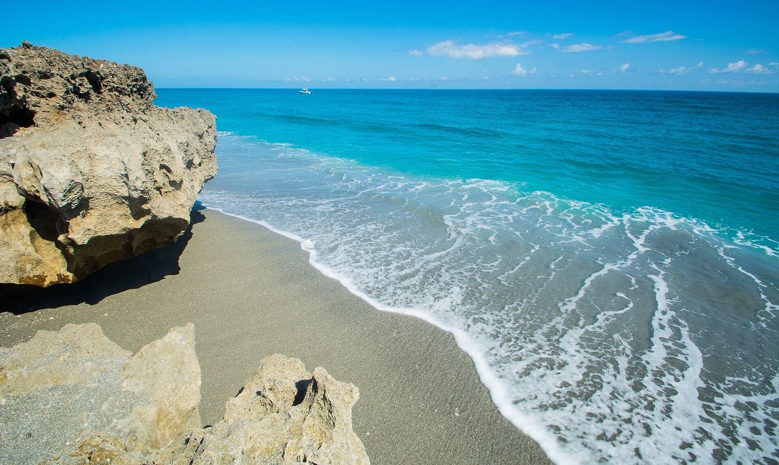 Turquoise surf washes across pale sand beside a rough limestone rock outcrop under a blue sky.