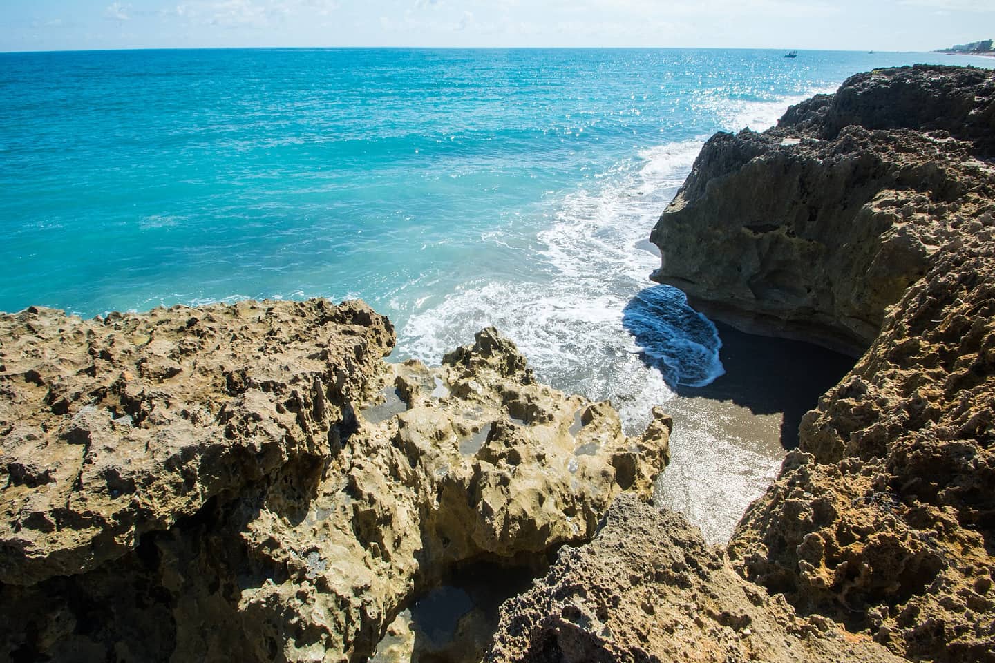 Rugged eroded limestone rocks frame a turquoise ocean wave breaking onto a dark sandy shoreline.