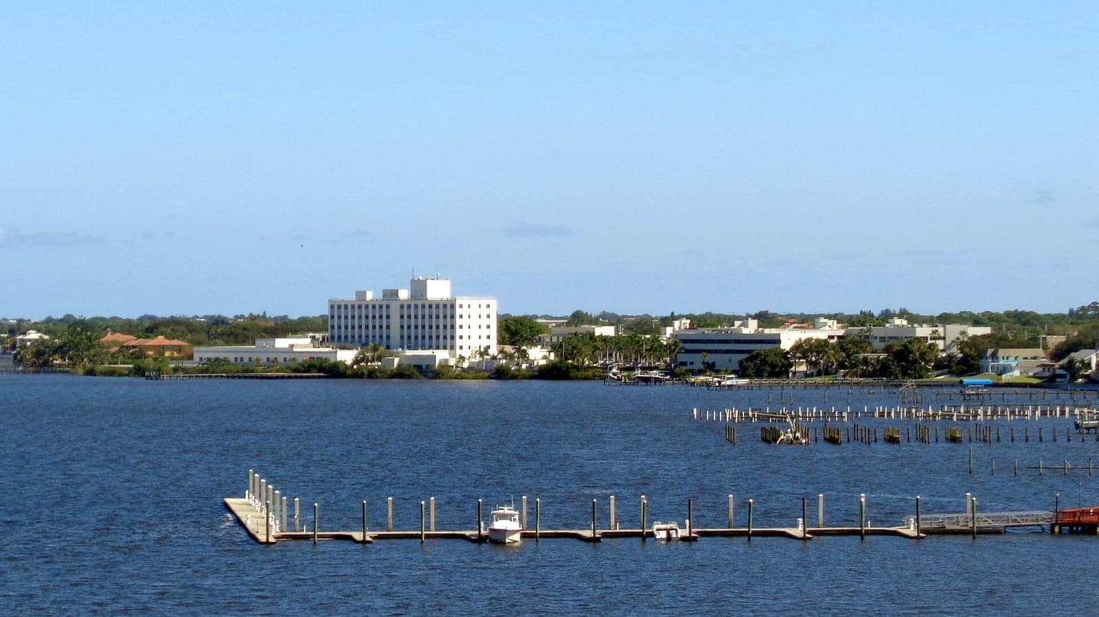 Calm river water with docks and a white multi-storey building across the shore under blue sky.